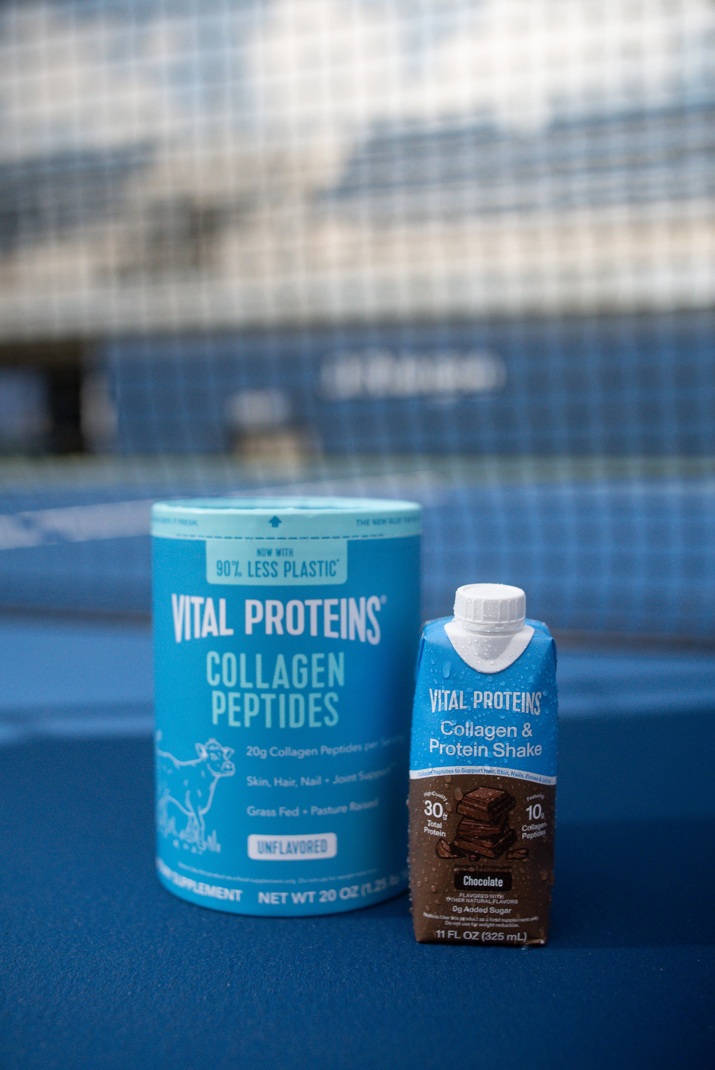 A container of Vital Proteins Collagen Peptides and a Vital Proteins Collagen & Protein Shake in chocolate flavor on a blue surface with a wire fence in the background.