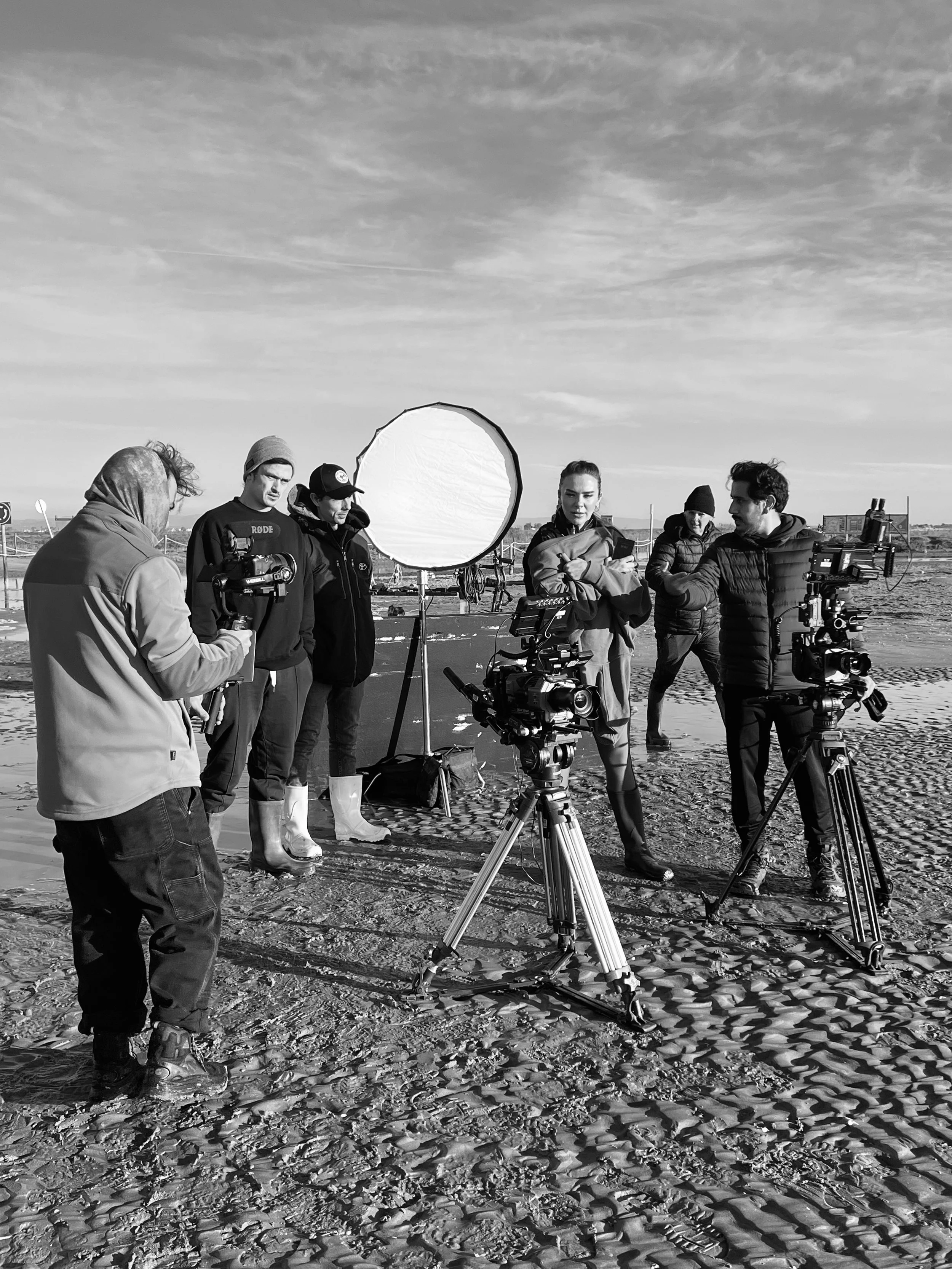 A film crew on a beach setting up equipment, including cameras and a large reflector, for a shoot in the early morning or late afternoon.