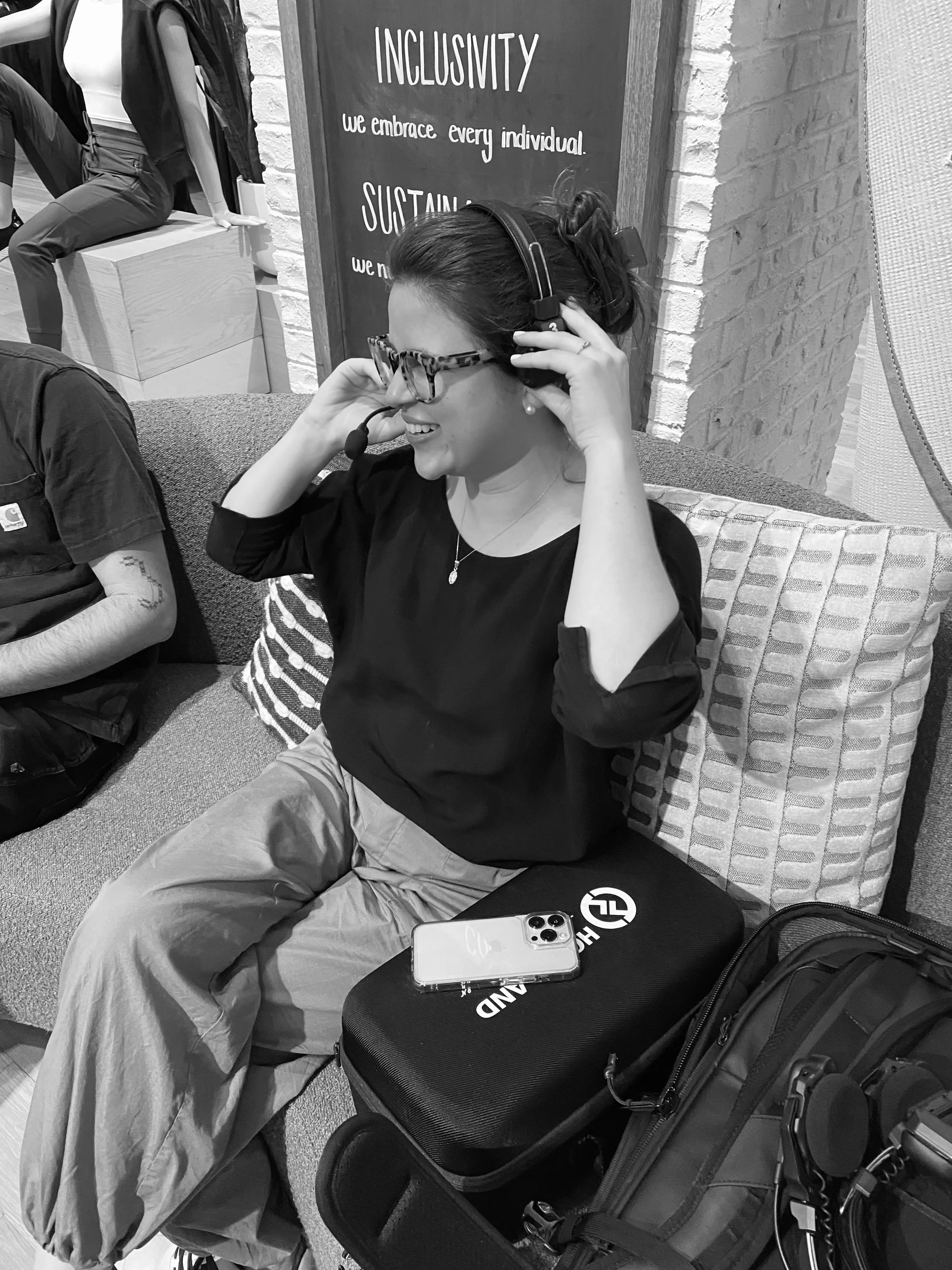 A woman wearing glasses and headphones sits on a couch, smiling, with a phone placed on a bag in front of her. There is a motivational sign on the wall behind her.