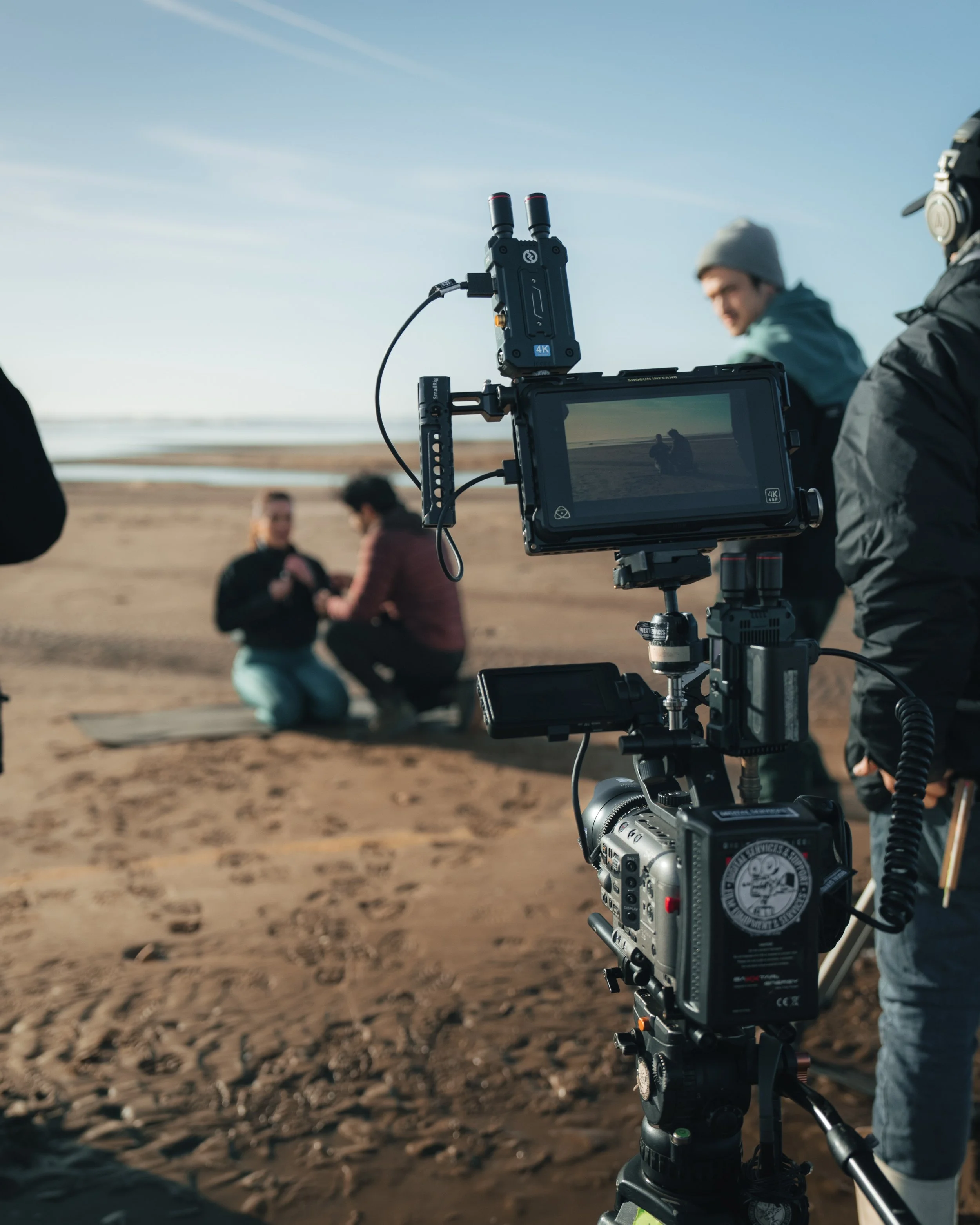 Film crew recording a scene on the beach with two people kneeling and talking, camera and crew equipment visible in the foreground.