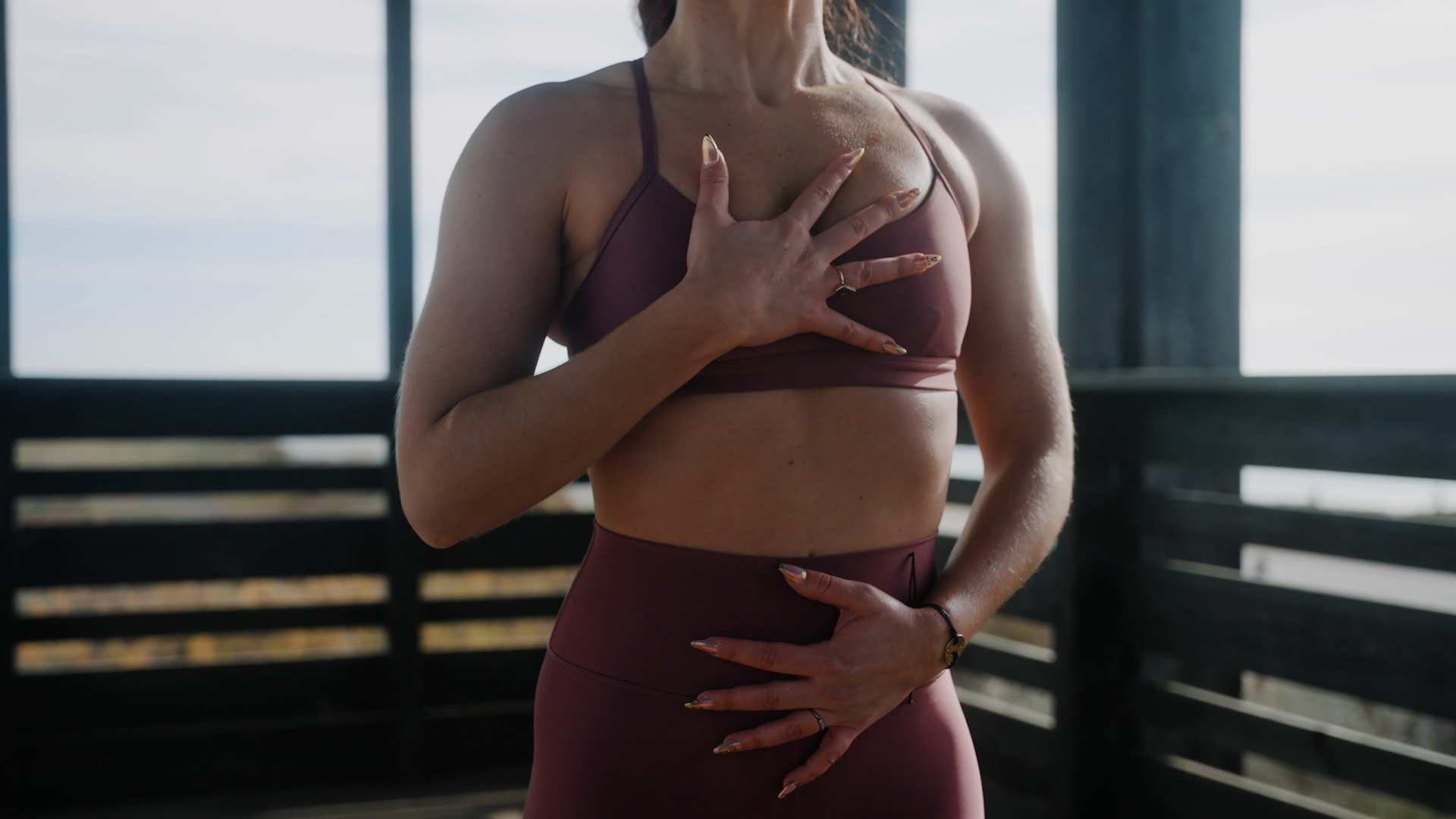 A woman in athletic wear practicing yoga in a wooden pavilion outdoors, with her hand placed over her chest.