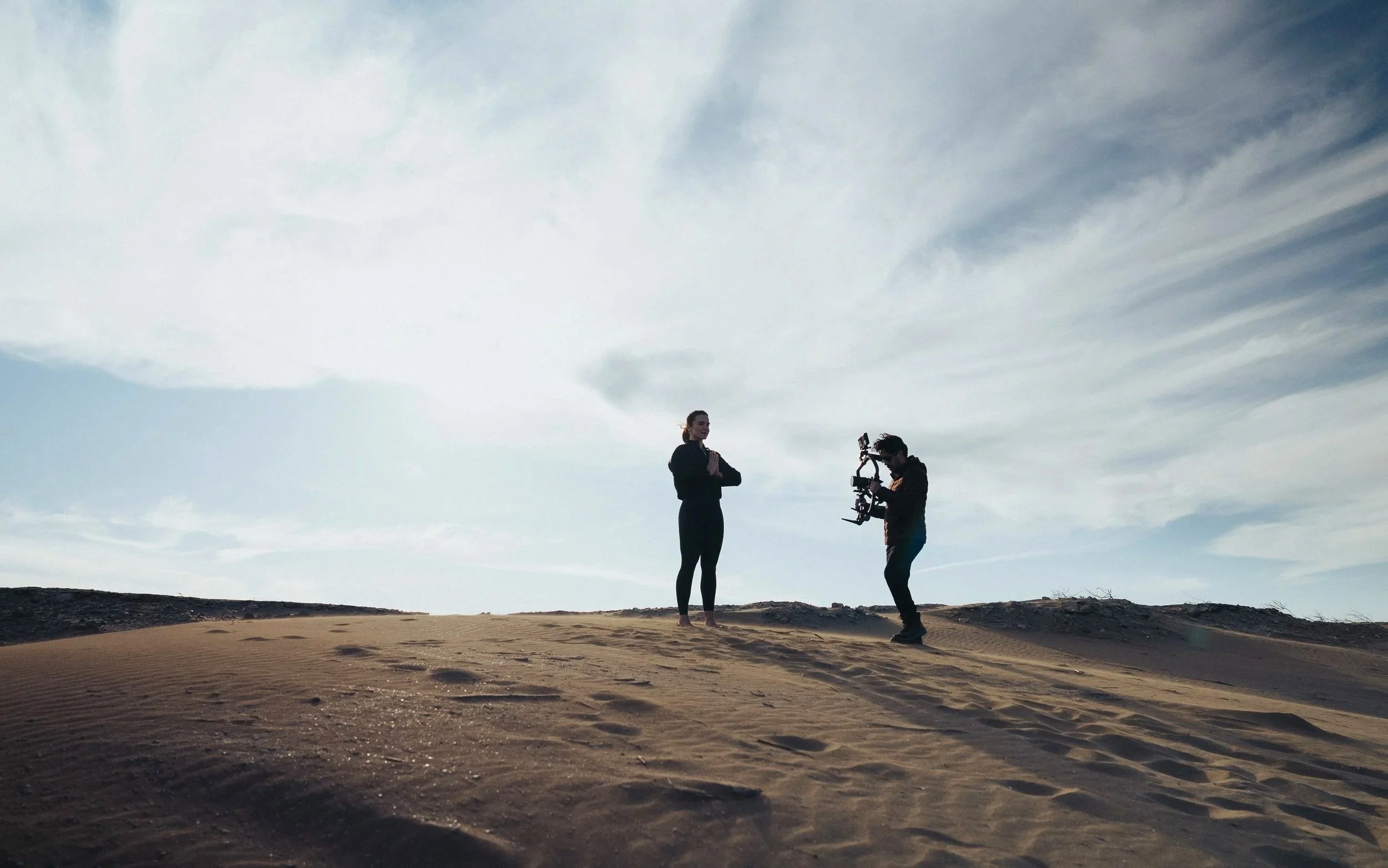 A person with a camera rig filming another person standing on sand dunes with a cloudy sky in the background.
