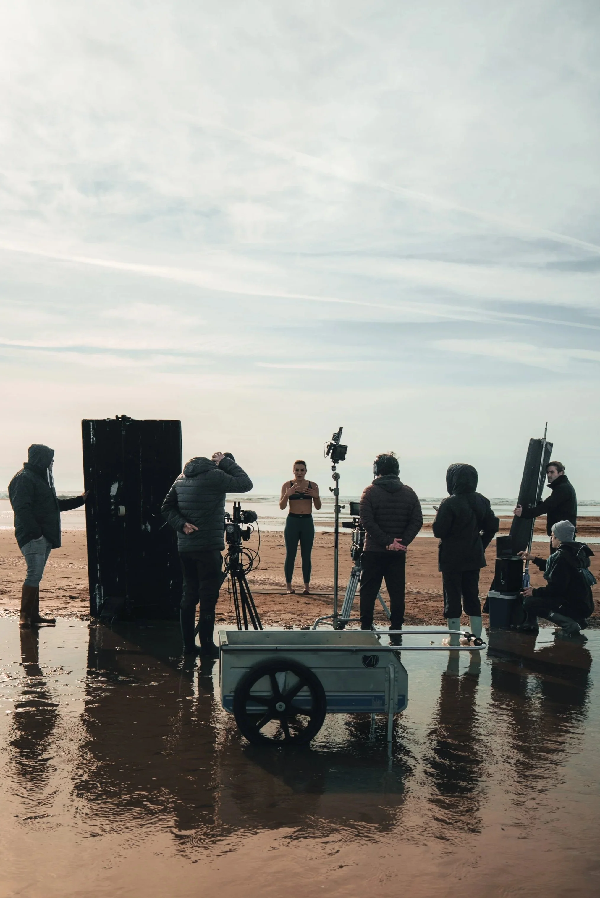 A film crew filming a woman on a beach with overcast sky, equipment including cameras, lighting, and reflectors present.