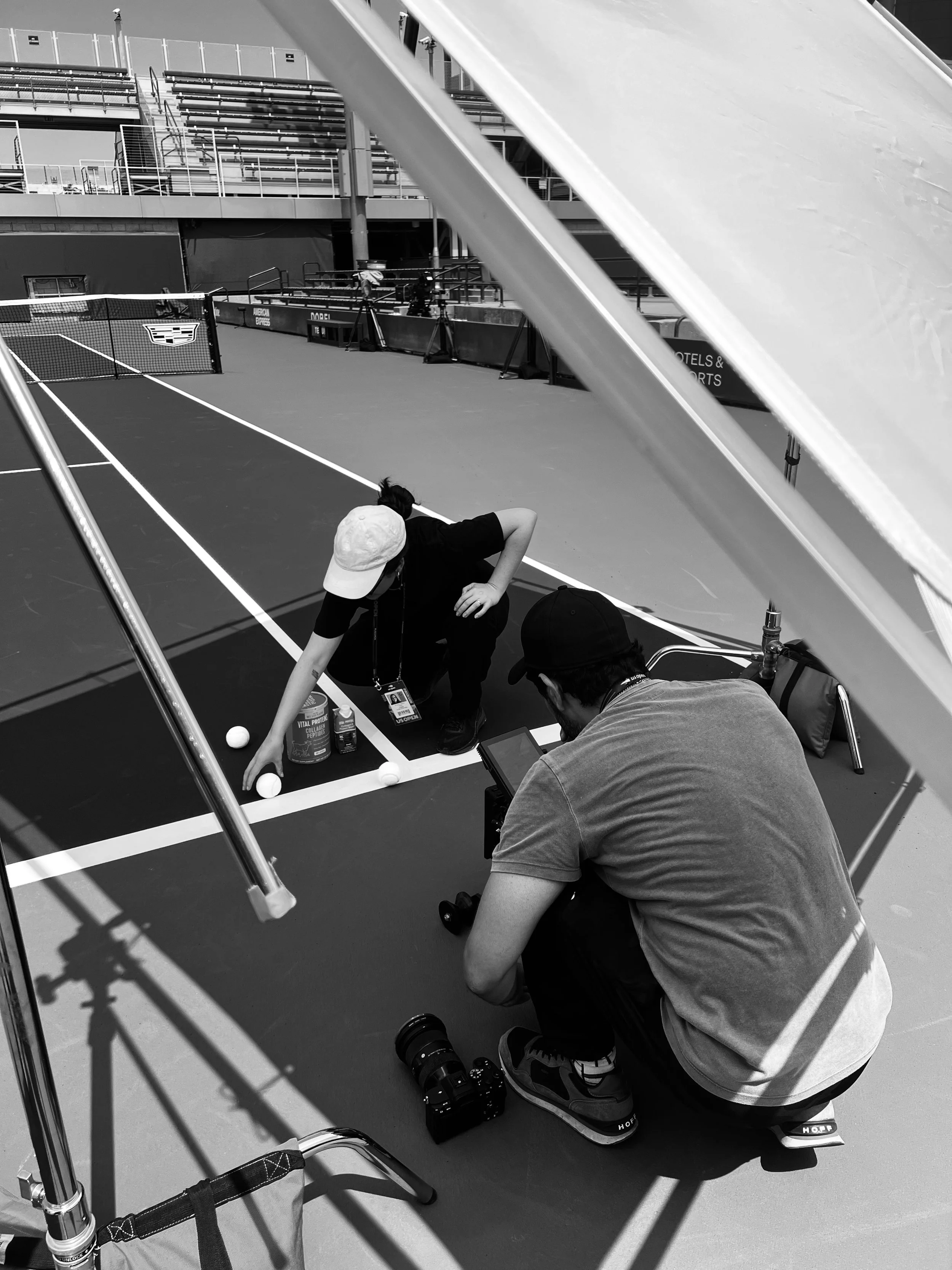 Photographers taking photos of a person setting up tennis balls on a tennis court, with camera equipment and supplies nearby.
