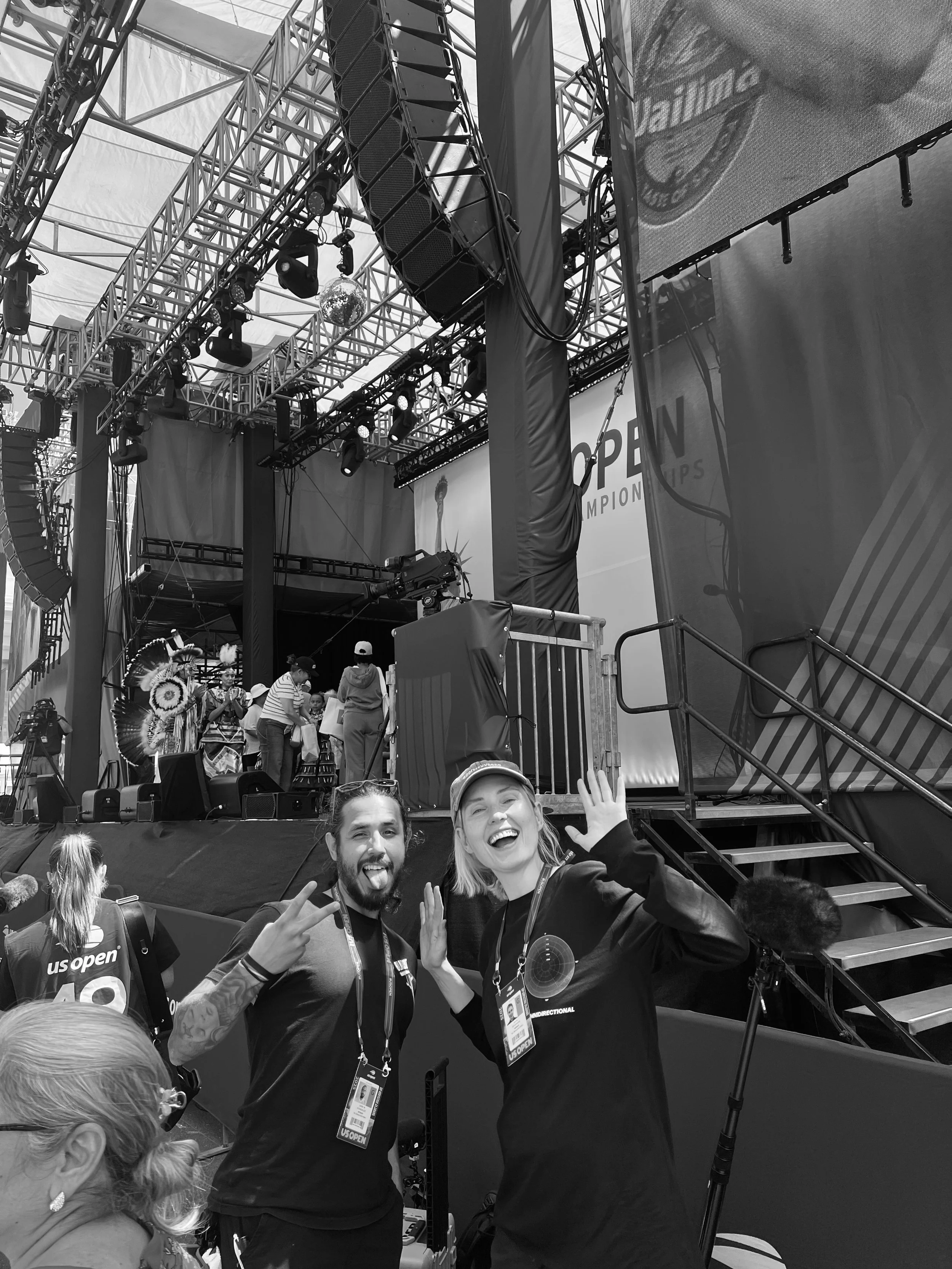 Two smiling people, a man and a woman, wearing US Open badges, pose for a photo at a tennis event. The man is making a peace sign, and the woman is waving. In the background, there is a stage with performers and large screens displaying the US Open logo.