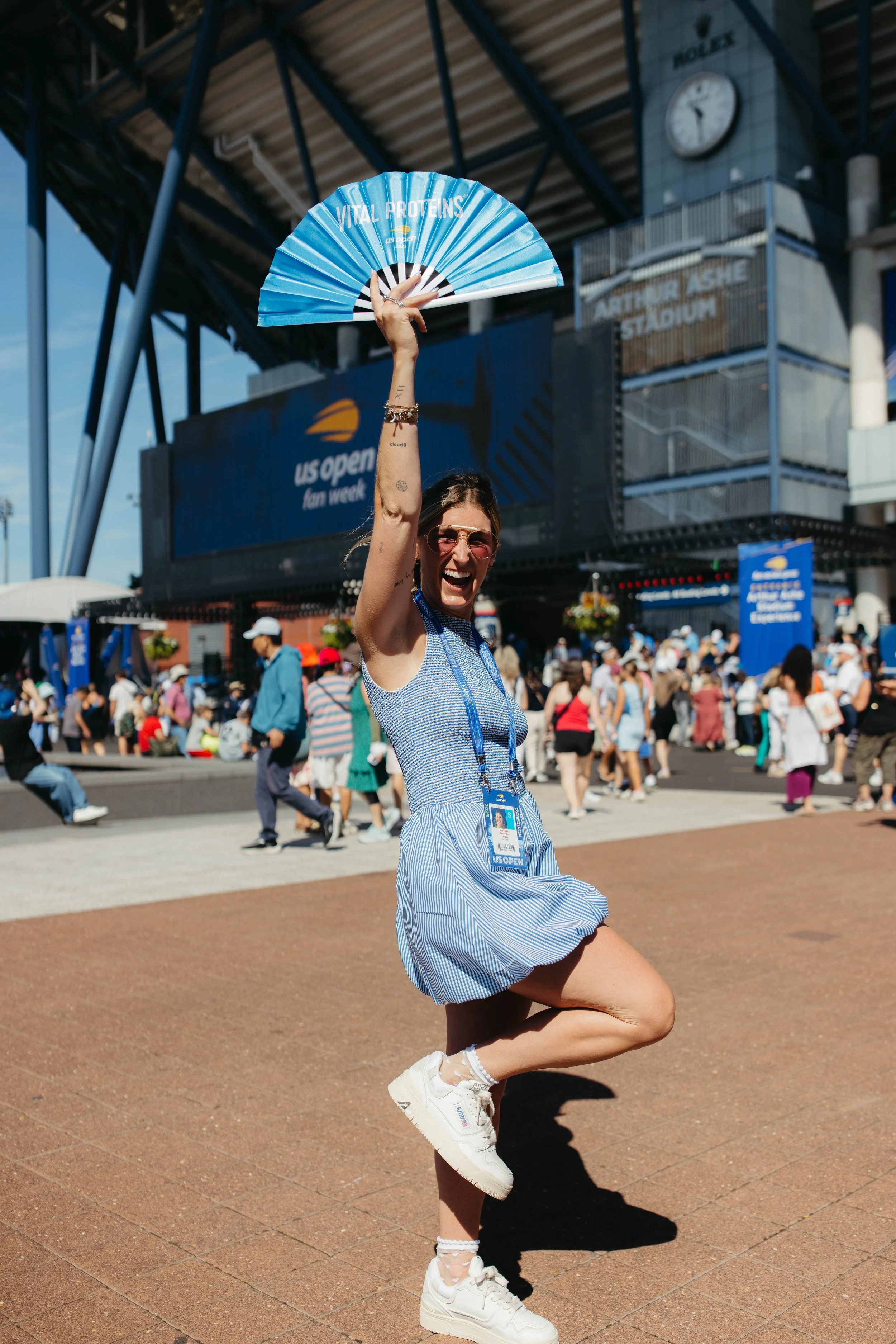 A woman in a blue dress and white sneakers celebrates at the US Open, holding a blue fan above her head, standing on a brick pathway at Arthur Ashe Stadium with a large crowd in the background.