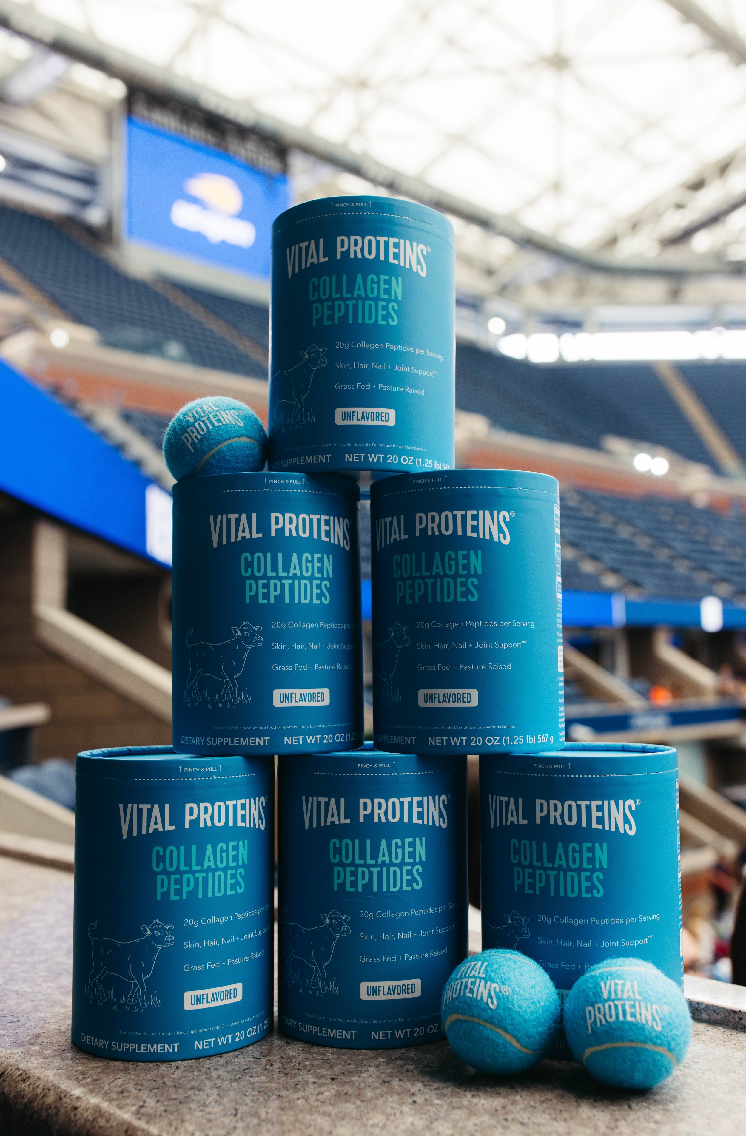 Multiple cans of Vital Proteins Collagen Peptides dietary supplement stacked on a surface with two tennis balls labeled Vital Proteins nearby, in an indoor sports arena.