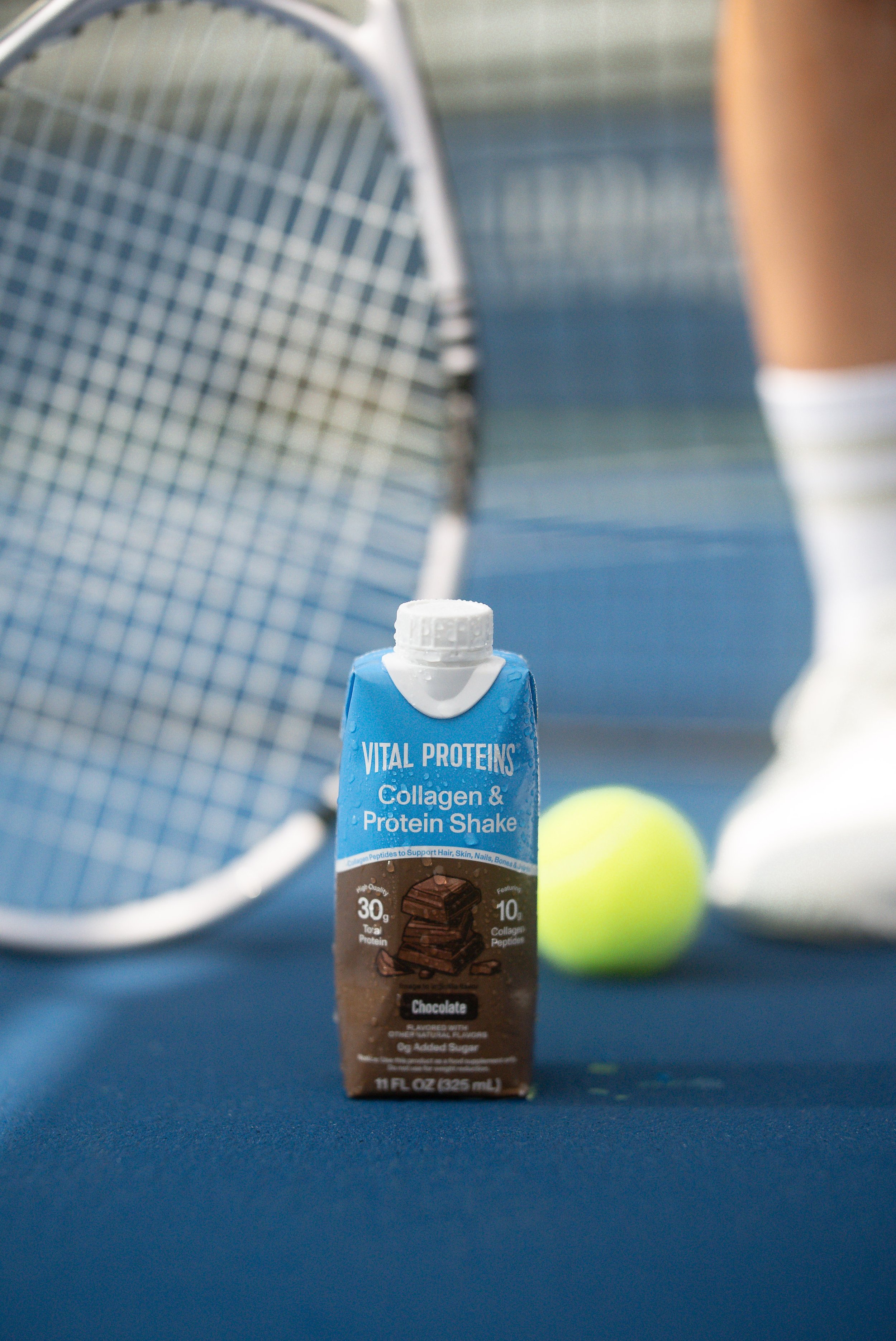 A carton of Vital Proteins Collagen & Protein Shake in chocolate flavor, and a tennis ball with a tennis racket in the background on a blue surface.