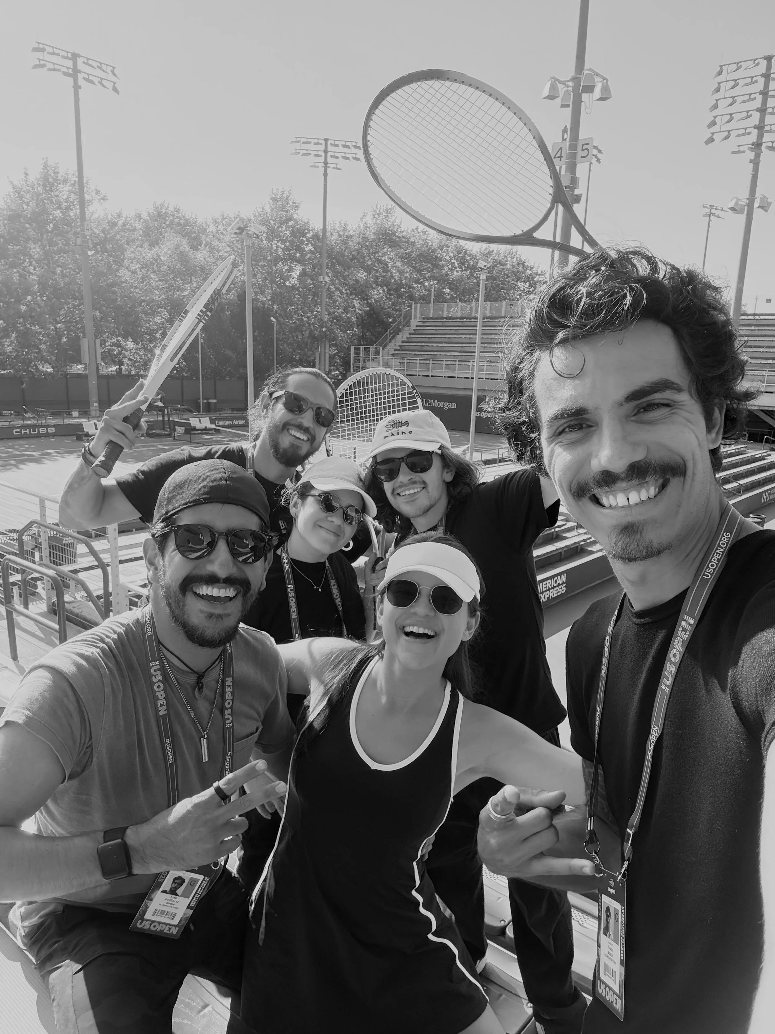 Group of five people taking a selfie on a tennis court, holding tennis rackets, with trees and empty bleachers in the background, all smiling and wearing sunglasses and casual athletic clothing.