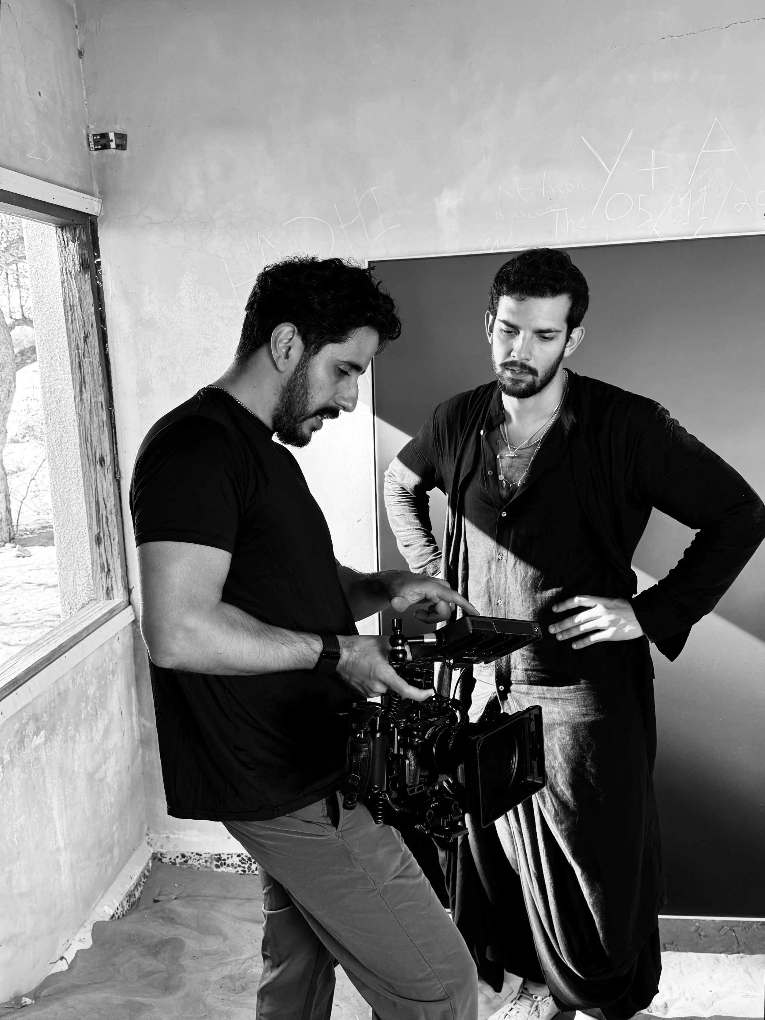 Two men with beards and dark hair review footage on a digital monitor on a film set. One man holds a professional camera, the other looks on with hands on hips.