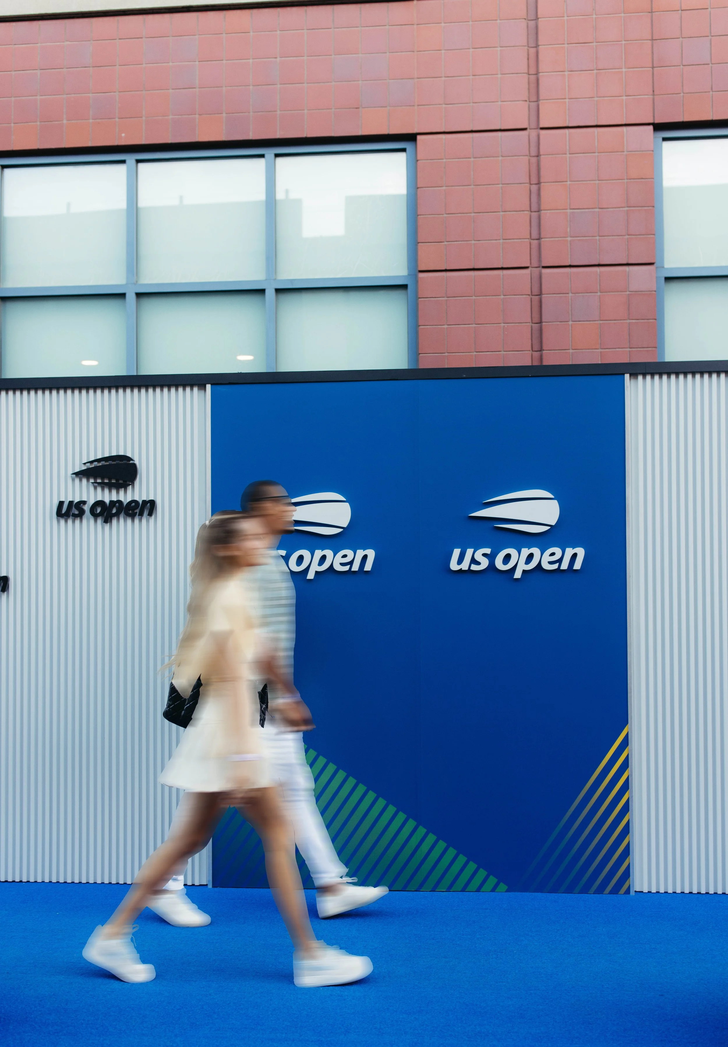 People walking past a blue sign at the US Open tennis tournament with the US Open logo, in front of a brick building.
