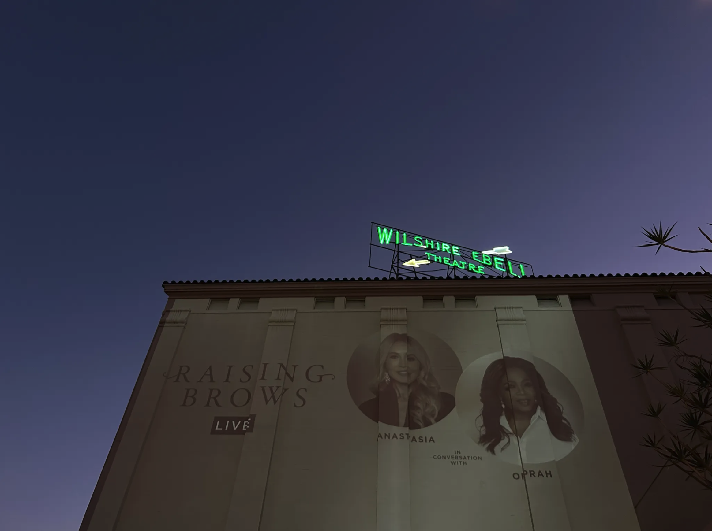 Nighttime view of the Wilshire Ebell Theatre with a green neon sign and a large poster for a live event called 'Raising Brows' featuring Anastasia and Oprah.