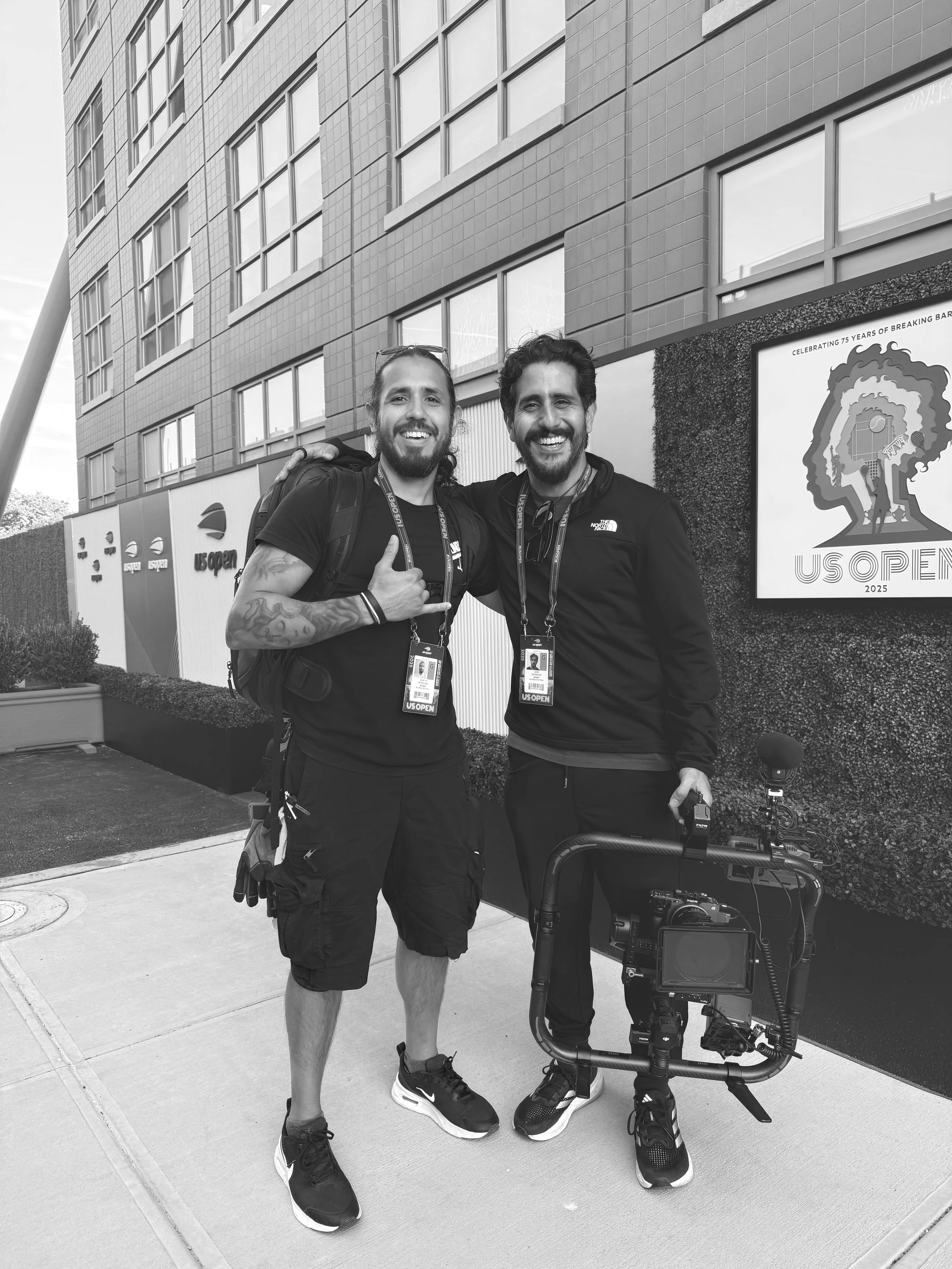 Two men smiling and posing for a photo outside a building with a sign that reads 'US Open'. One man is holding a camera stabilizer, and both are wearing event badges around their necks.