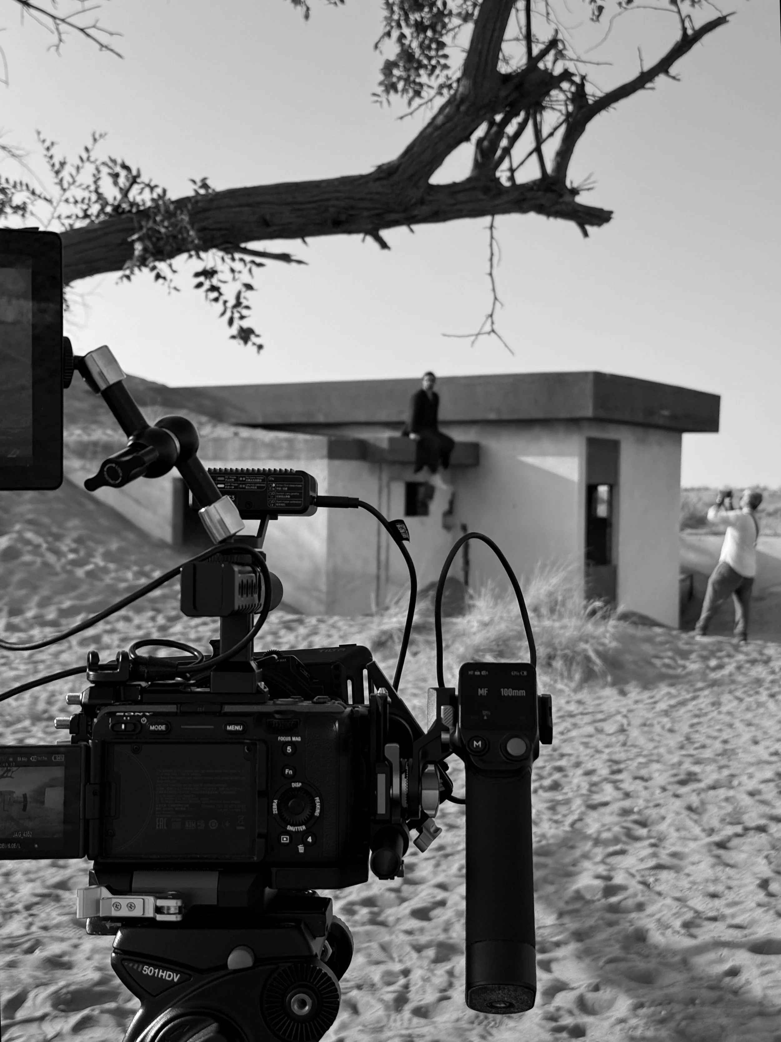 Black and white photo of a landscape scene with a camera setup in the foreground, focusing on a small, modern house with a woman sitting on the rooftop and a person walking nearby. There are trees and sandy terrain around the scene.