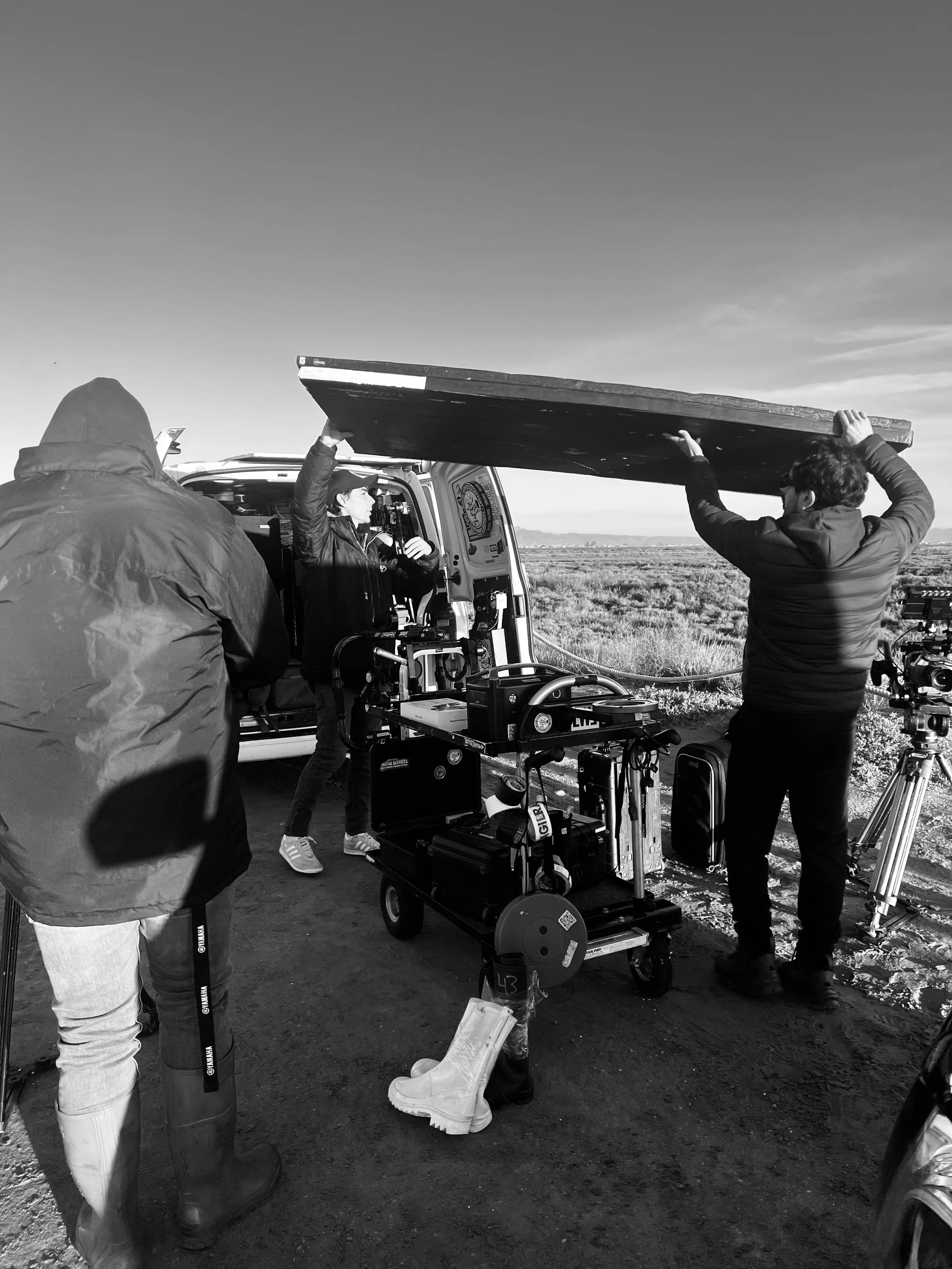 Film crew setting up equipment outdoors near a van, with one person holding a large lighting reflector and another standing nearby, while a person operates a camera on a tripod in a vast open field.