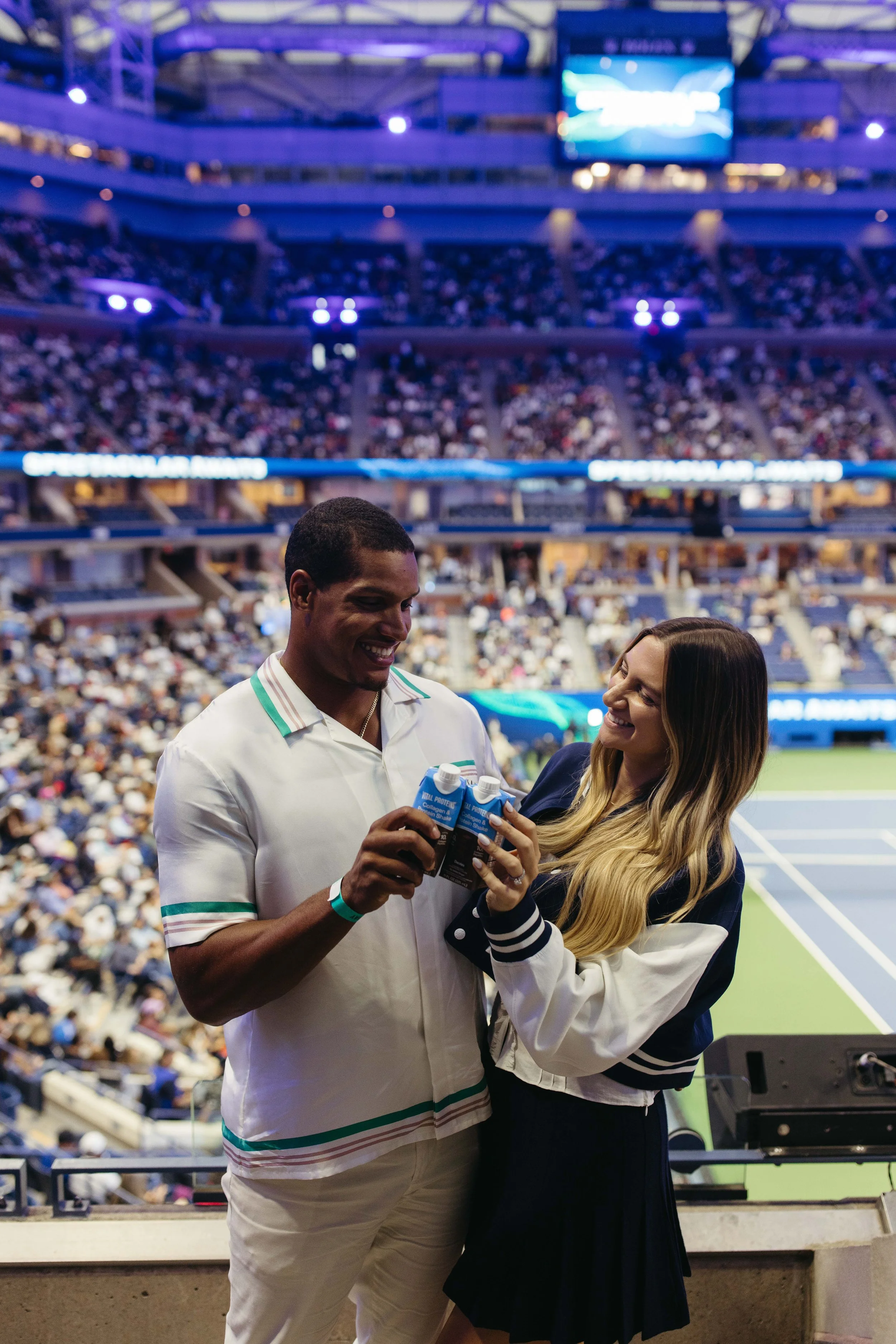 A man and woman smiling and holding bottles of water at a tennis stadium, with a large crowd in the background.