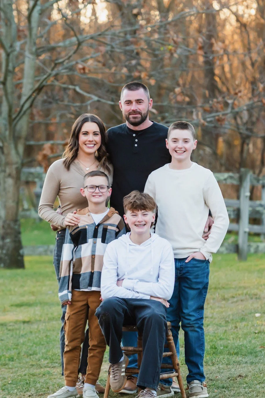 A family of six posing outdoors during fall, with trees in the background. The family includes a woman, a man, and four boys, all smiling at the camera.