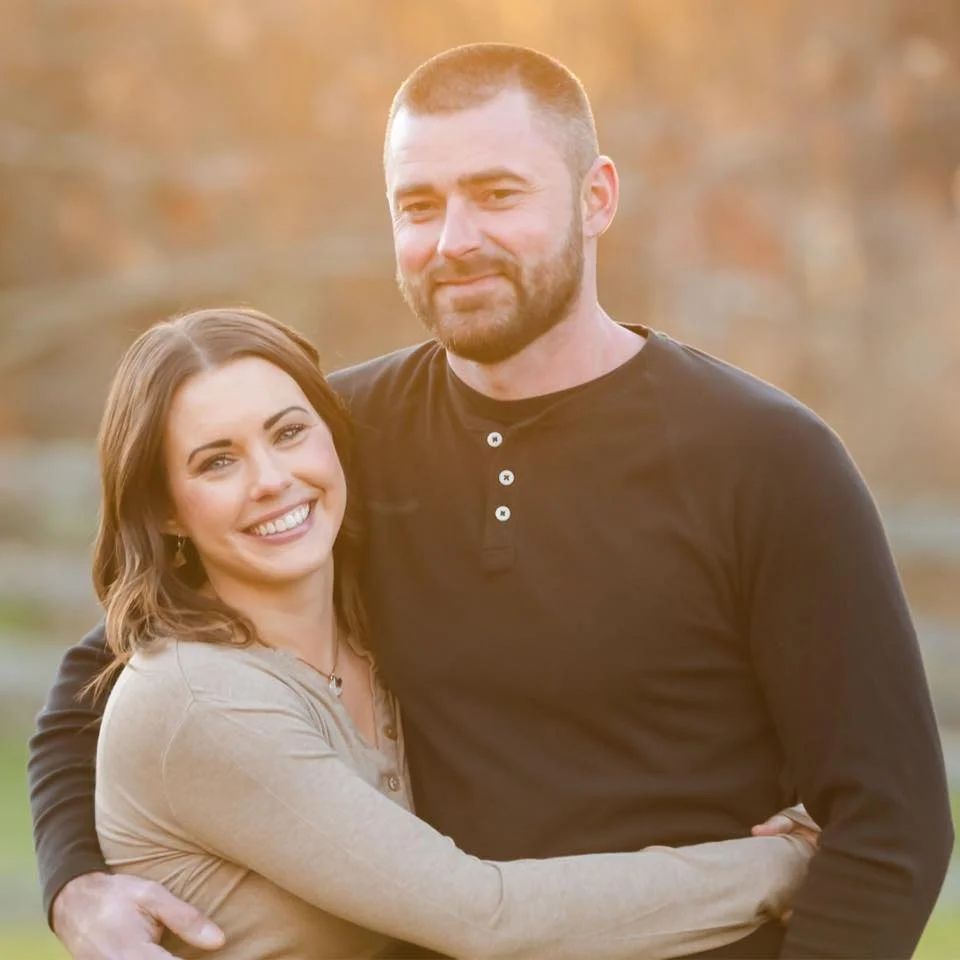 A smiling woman with shoulder-length brown hair hugging a bearded man with short hair, outdoors in warm sunlight with autumn-colored trees in the background.