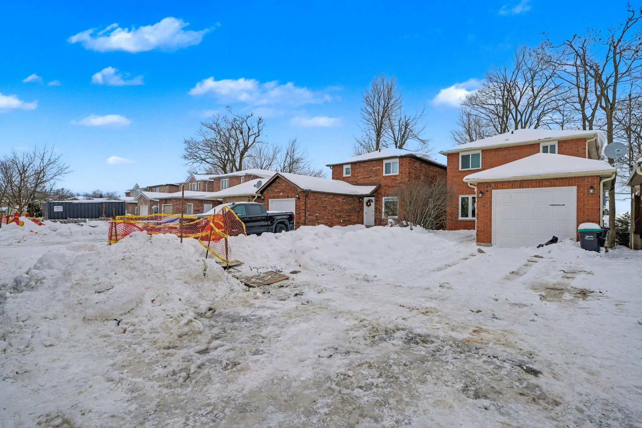 Snow-covered residential neighborhood with brick houses, leafless trees, parked vehicles, and a bright blue sky with some clouds.