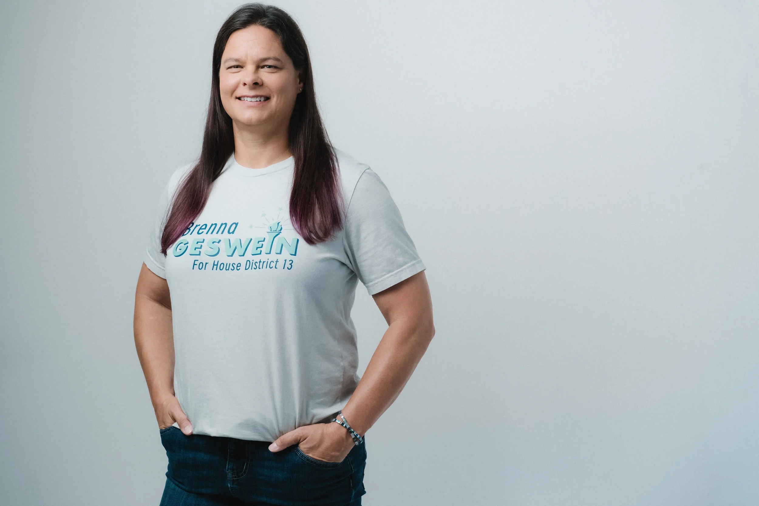 A woman standing with her left hand in her pocket, smiling, wearing a light gray t-shirt with blue text that reads 'Brenna Geswein For House District 13' against a plain light gray background.