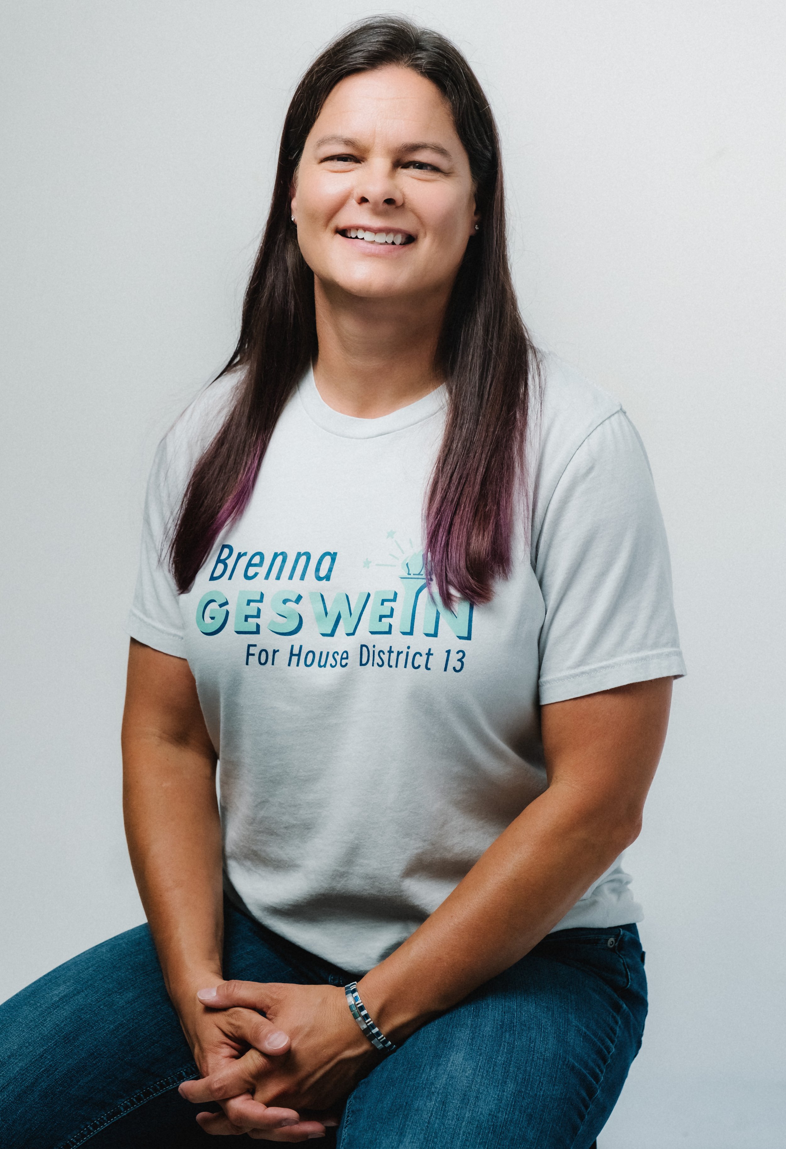 Portrait of a smiling woman with long dark hair, wearing a gray campaign T-shirt, sitting with legs crossed against a plain white background.
