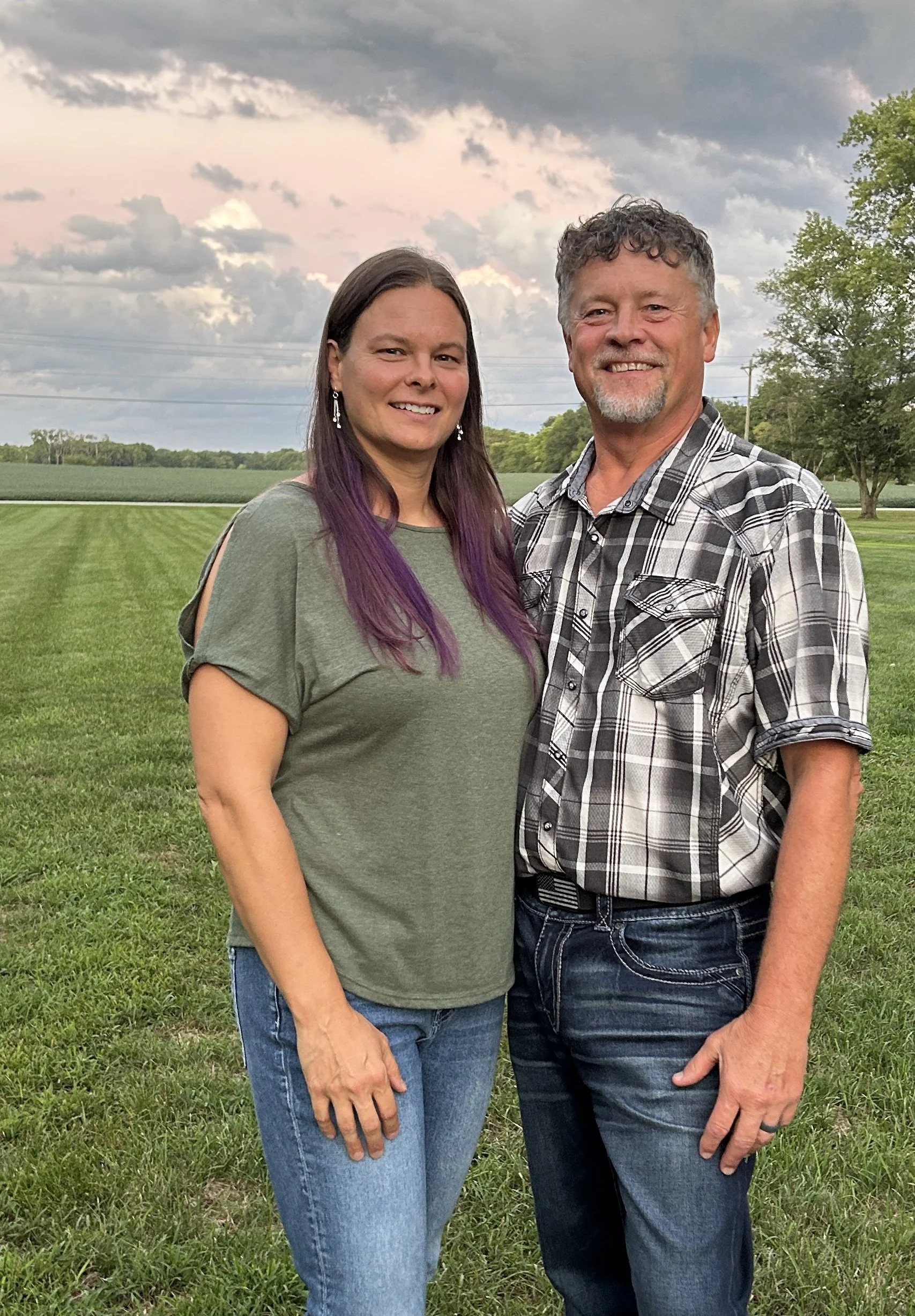 A woman and a man standing close together outdoors in a grassy field with trees and a cloudy sky in the background, smiling at the camera.
