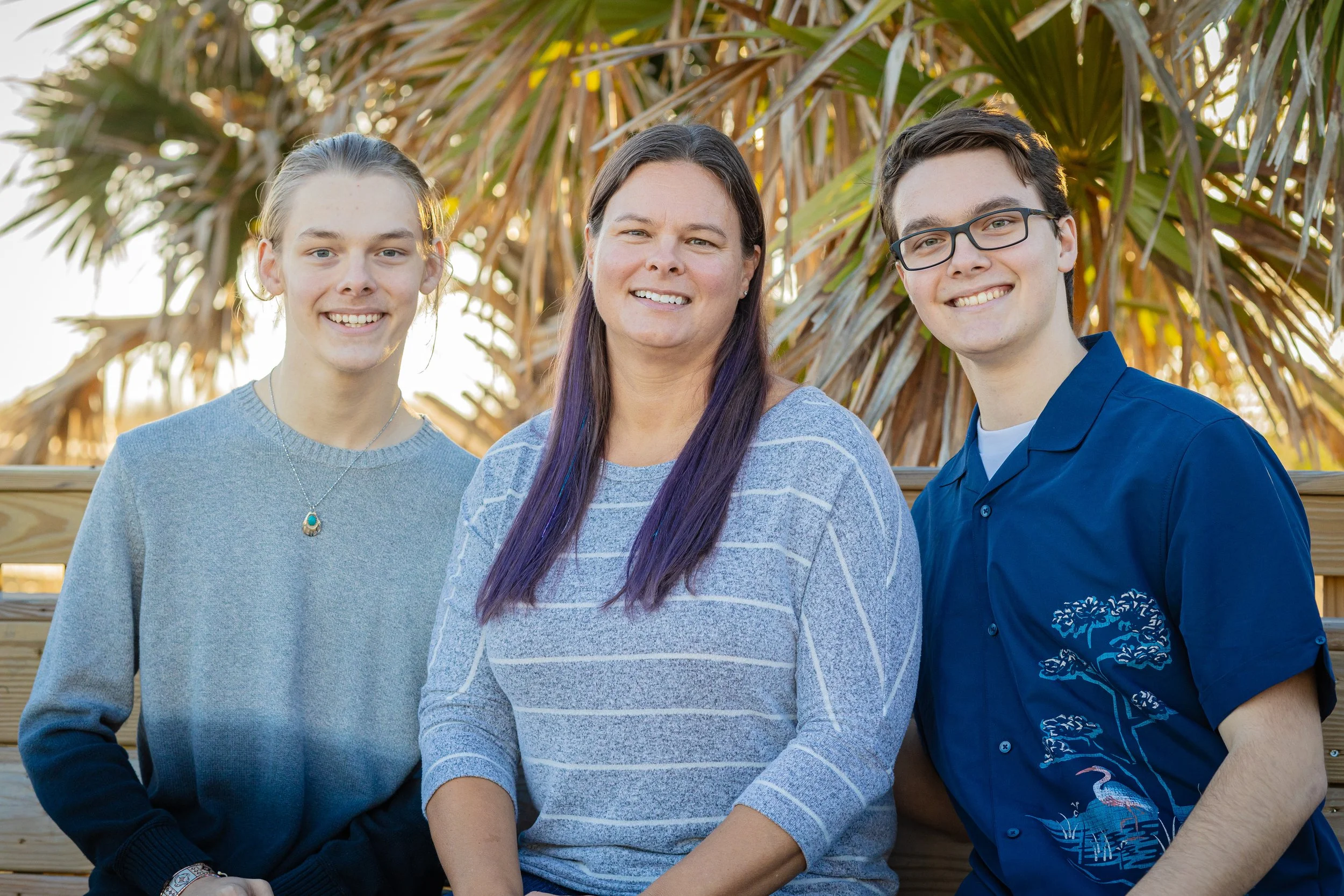 A woman with long purple hair and two teenagers, standing outdoors in front of palm trees, smiling.