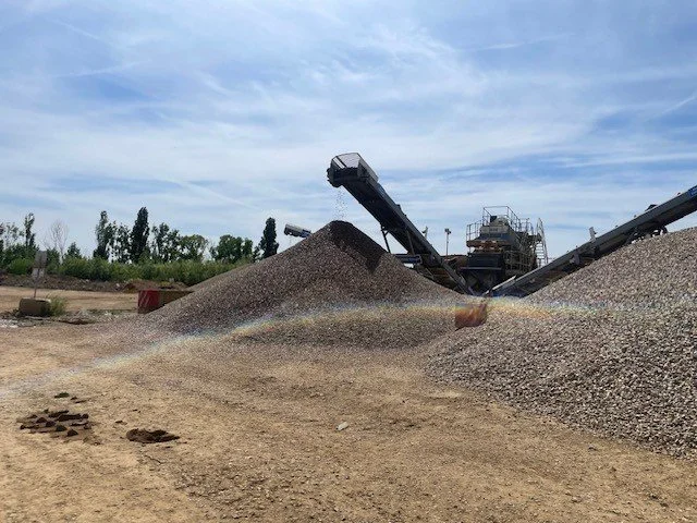 A gravel processing plant with conveyor belts transferring gravel into large piles on a dirt ground, under a blue sky with some clouds.