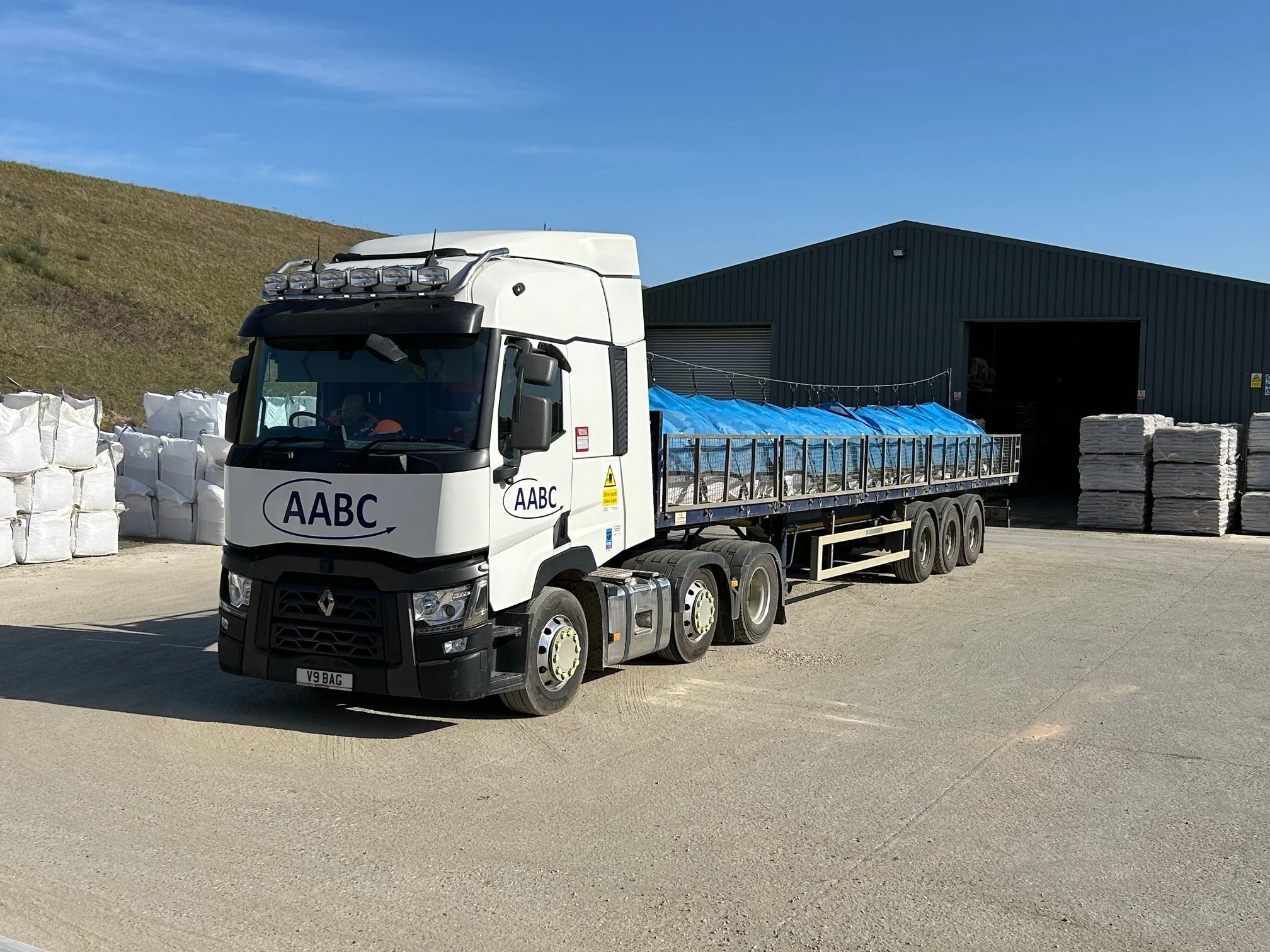 A white flatbed truck with the logo 'AABC' on the front and side, parked outside a large storage building. The truck's cargo area is loaded with covered items under blue tarps. Behind the truck, there are stacks of white wrapped bags and pallets of white bags near the building.