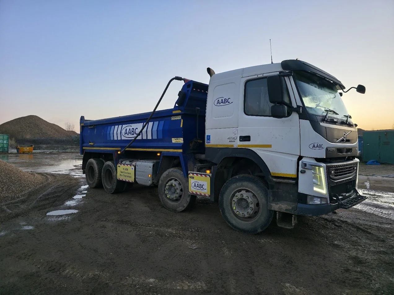 A white and blue dump truck with the company logo 'AABC' parked on a muddy construction site at sunset.