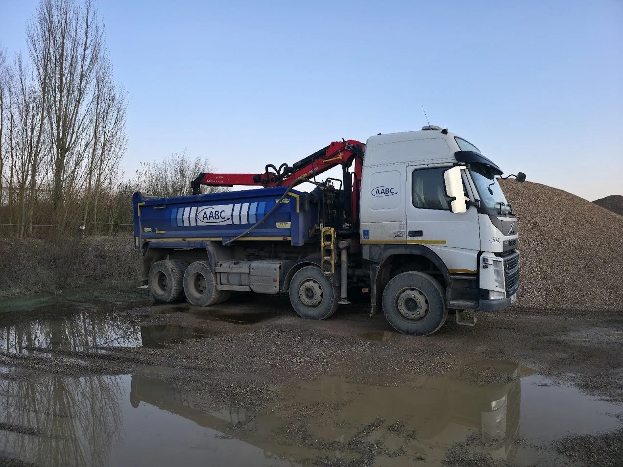 A white and blue construction truck with a red crane parked on a gravel surface near a large pile of gravel, with muddy puddles in the foreground and leafless trees in the background.