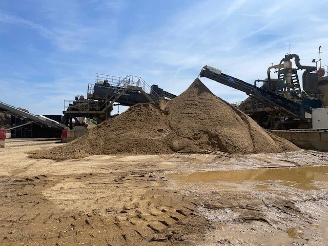 A large pile of sand on an industrial site with conveyor belts and machinery in the background.