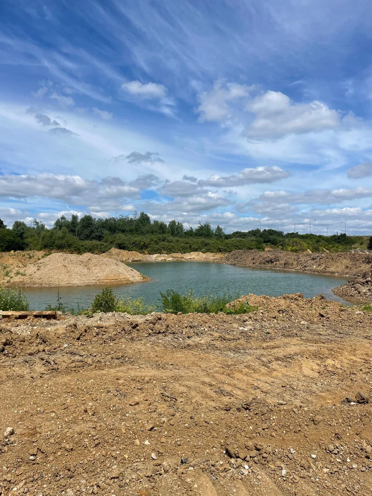 A construction site with a small body of water surrounded by dirt and rocks, with green bushes and trees in the background under a partly cloudy blue sky.