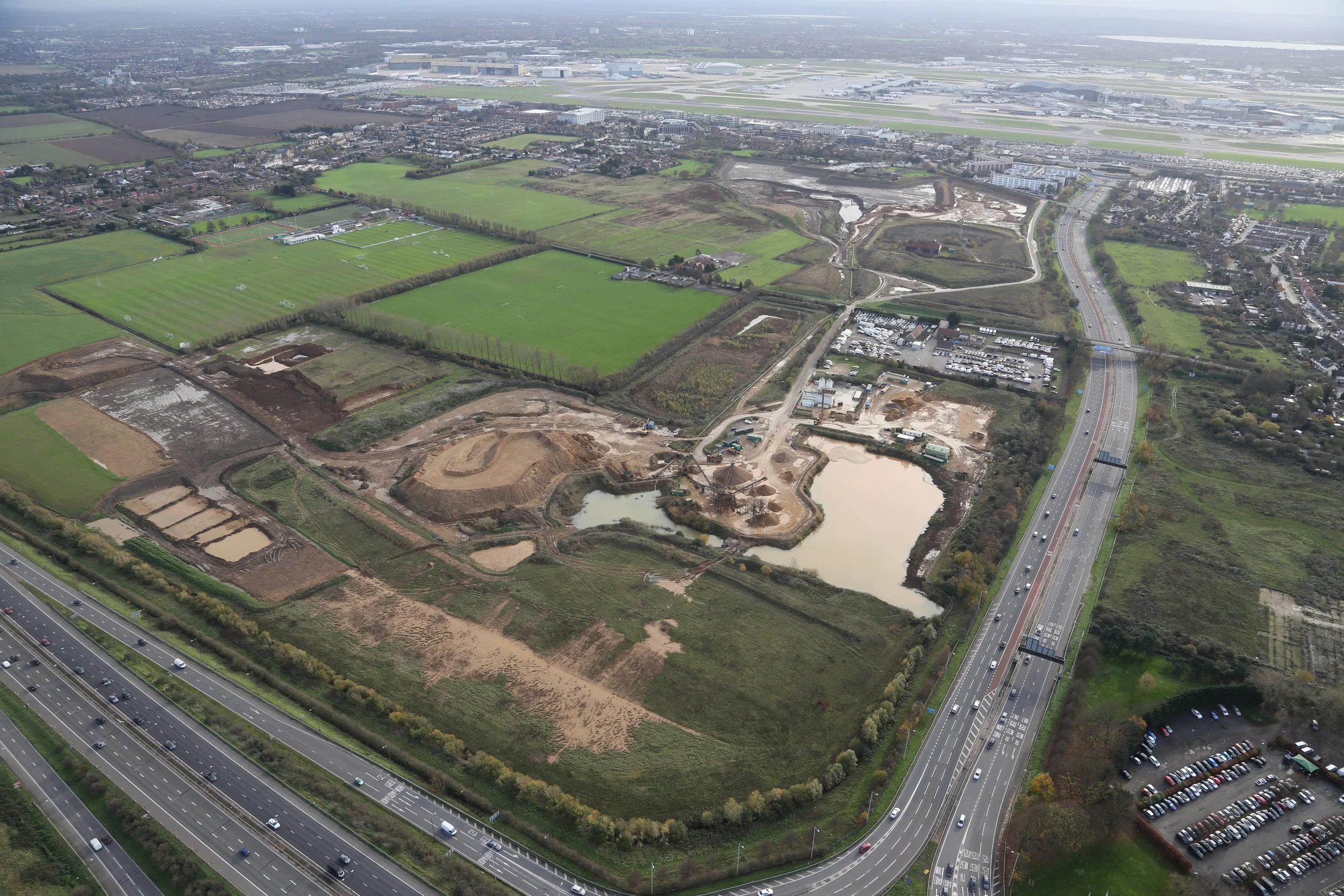 Aerial view of a construction site with dirt and water, adjacent to a highway, with green fields, residential areas, and airport runways in the background.