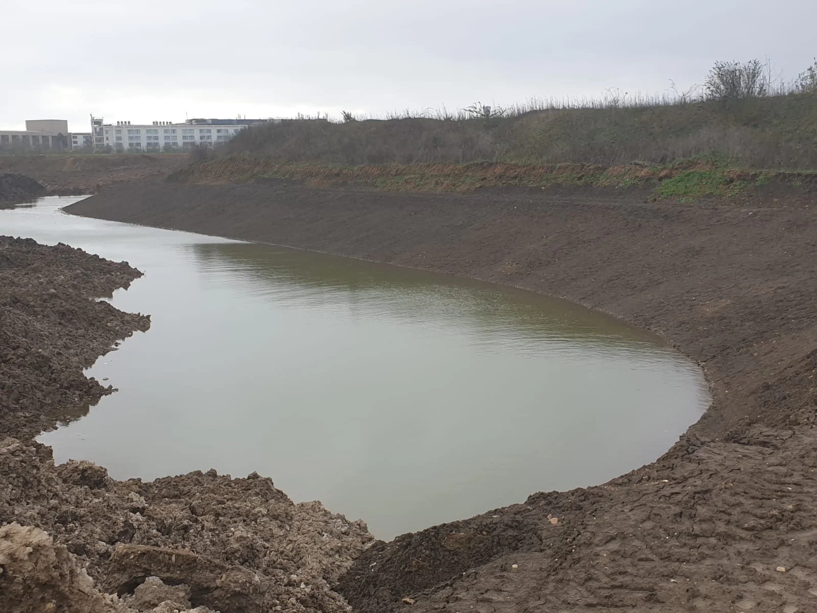 A partially excavated area with a small, curved body of water and exposed dirt banks, with buildings in the background under an overcast sky.