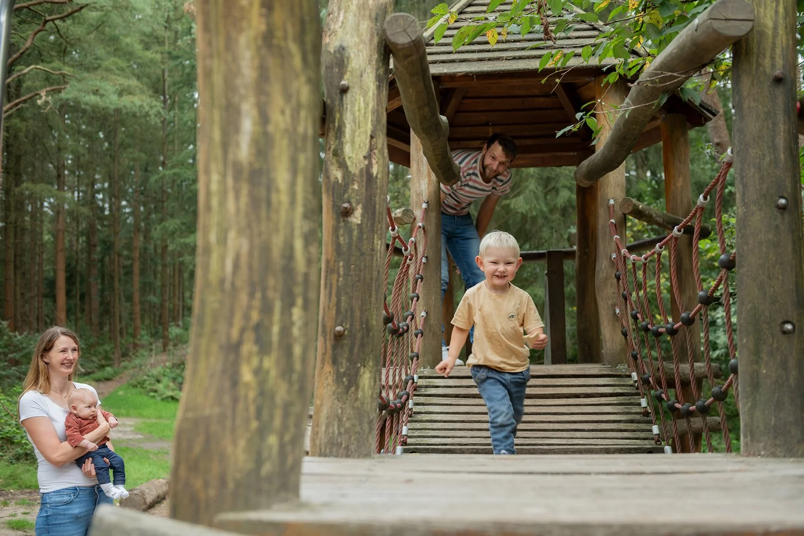 Familie im Wald beim Spielen auf einer Holzbank, die wie eine Baumhütte gestaltet ist, mit einem kleinen Jungen, der die Leiter herunterläuft, und einer Frau, die ein Baby hält, beide lachen und haben Spaß.
