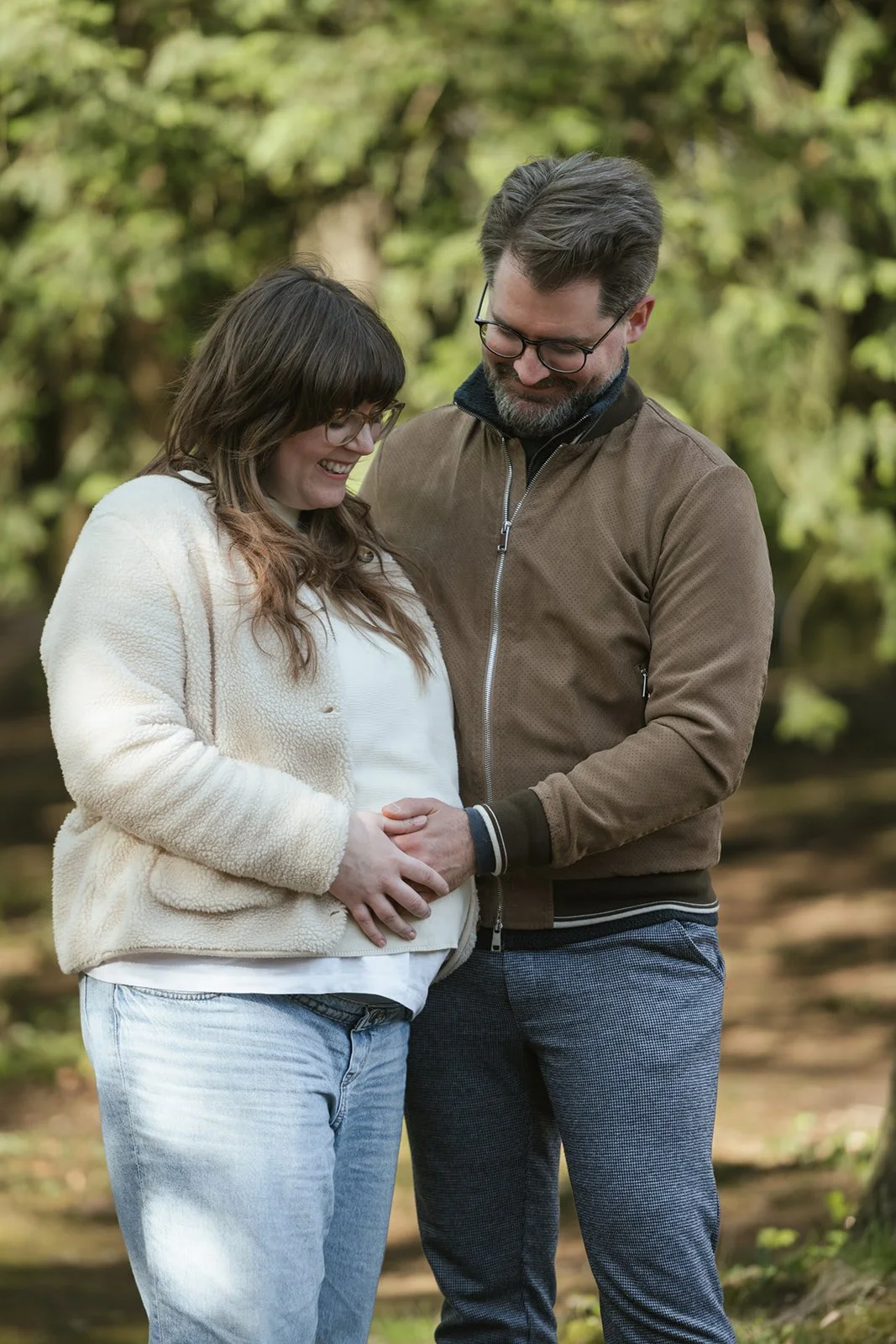 Ein Paar steht in einer Naturumgebung, lächelt und schaut liebevoll auf den Bauch. Die Frau ist schwanger und hat Babybauch.