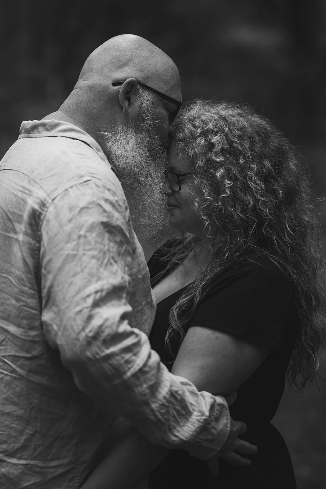 Ein Mann mit Brille und Vollbart und eine Frau mit Locken, die sich berühren, sich mit den Stirnen und Nasen berühren, in einer emotionalen Umarmung, schwarz-weiß Fotografie