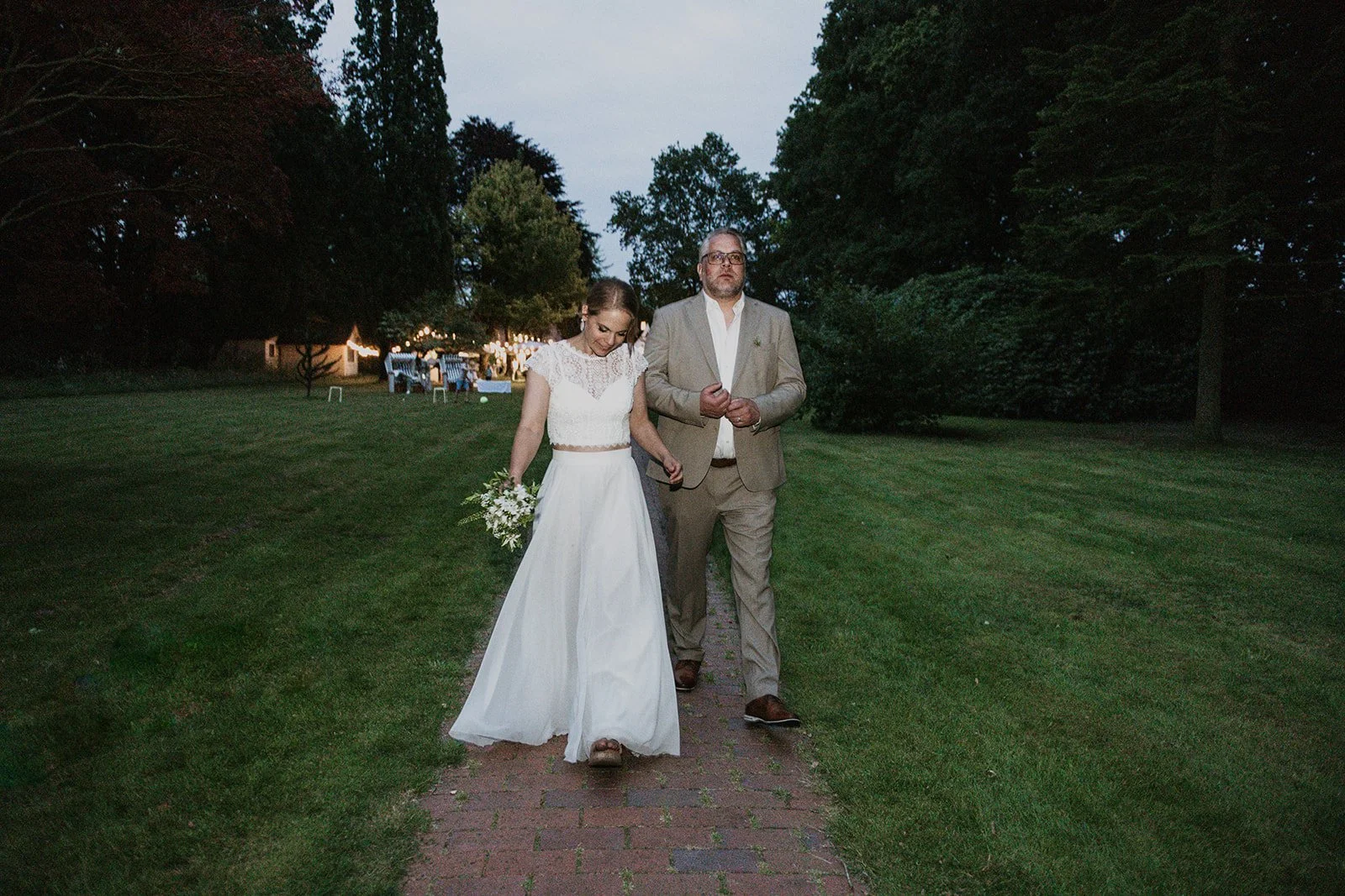 Brautpar laufen zum See, im Hintergrund ist die Hochzeitsparty im Sonnenuntergang mit Lichter im Park zu sehen.