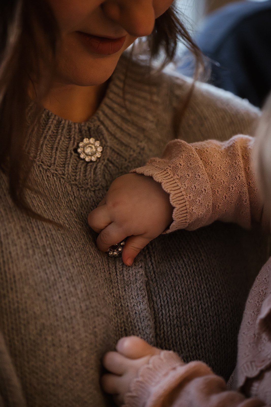 Nahaufnahme einer Frau, die ein Kind umarmt, nur ihre Lippen, Nase und Hände sind sichtbar, sie trägt einen braunen Pullover mit einer Schmuckbrosche, das Kind trägt eine rosa Strickjacke mit Spitzenbündchen.