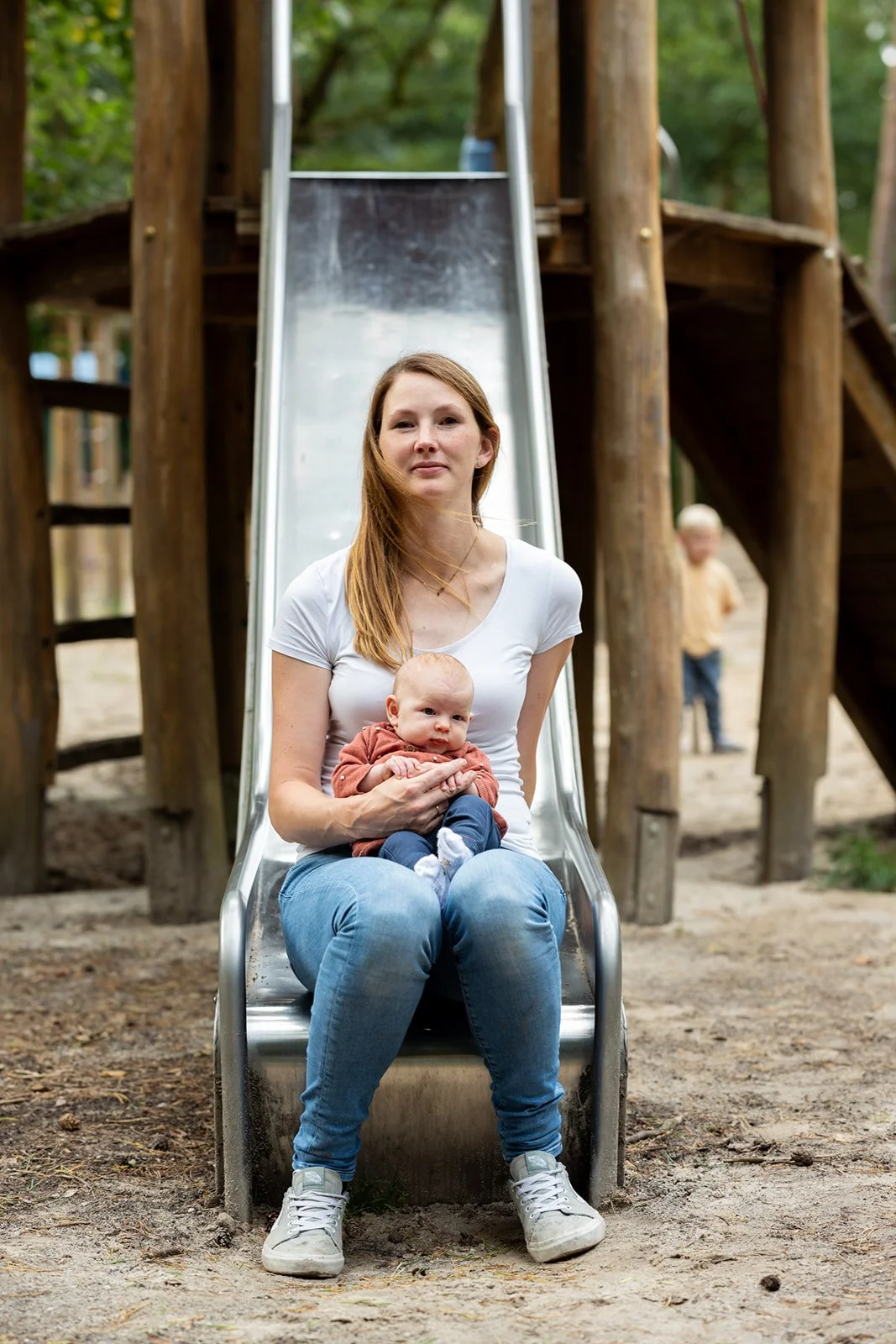 Eine Frau sitzt auf einer Rutsche in einem Spielpark, hält ein Baby im Arm, während ein Junge im Hintergrund im Sand spielt.
