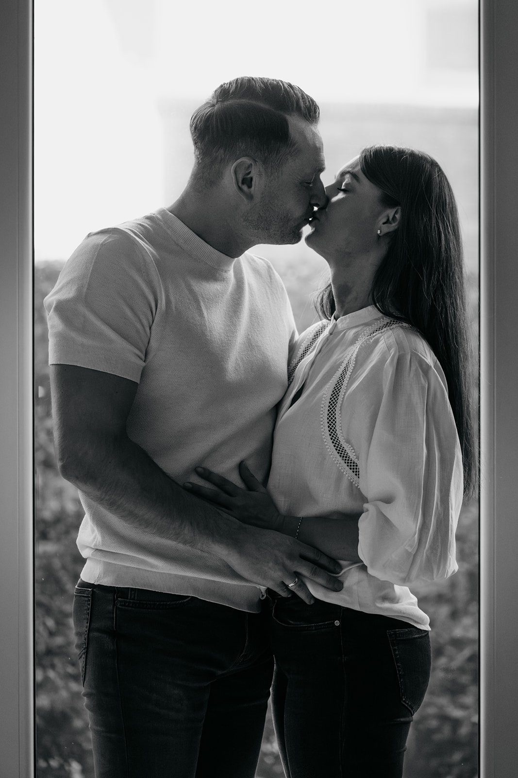 Paar, das sich küsst, vor dem Fenster mit Blick auf die Natur im Hintergrund, schwarz-weiß Foto.