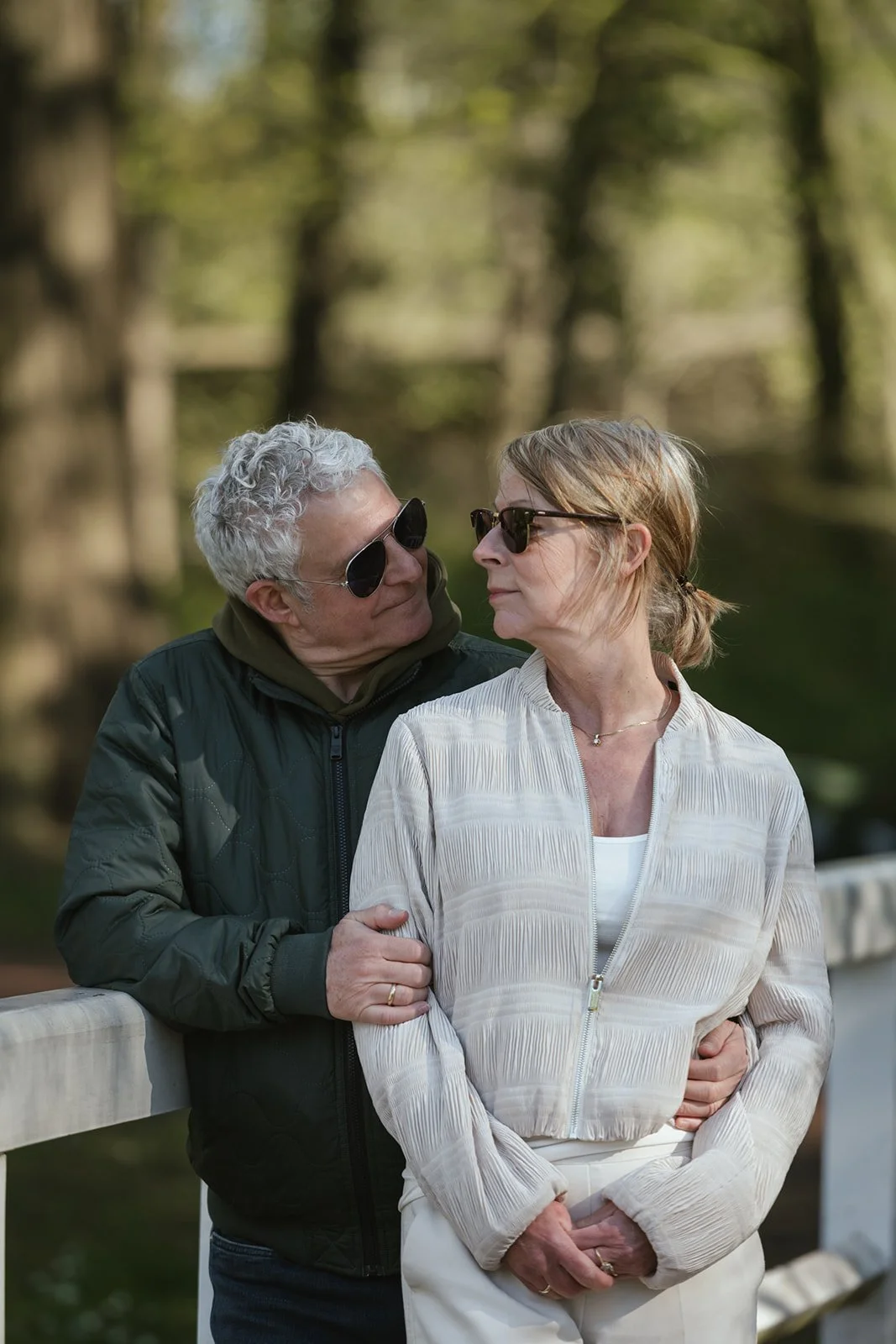 Ein älteres Paar steht eng beieinander auf einer Brücke im Freien, beide tragen Sonnenbrillen und schauen sich tief in die Augen. Im Hintergrund sind Bäume und Wasser sichtbar.