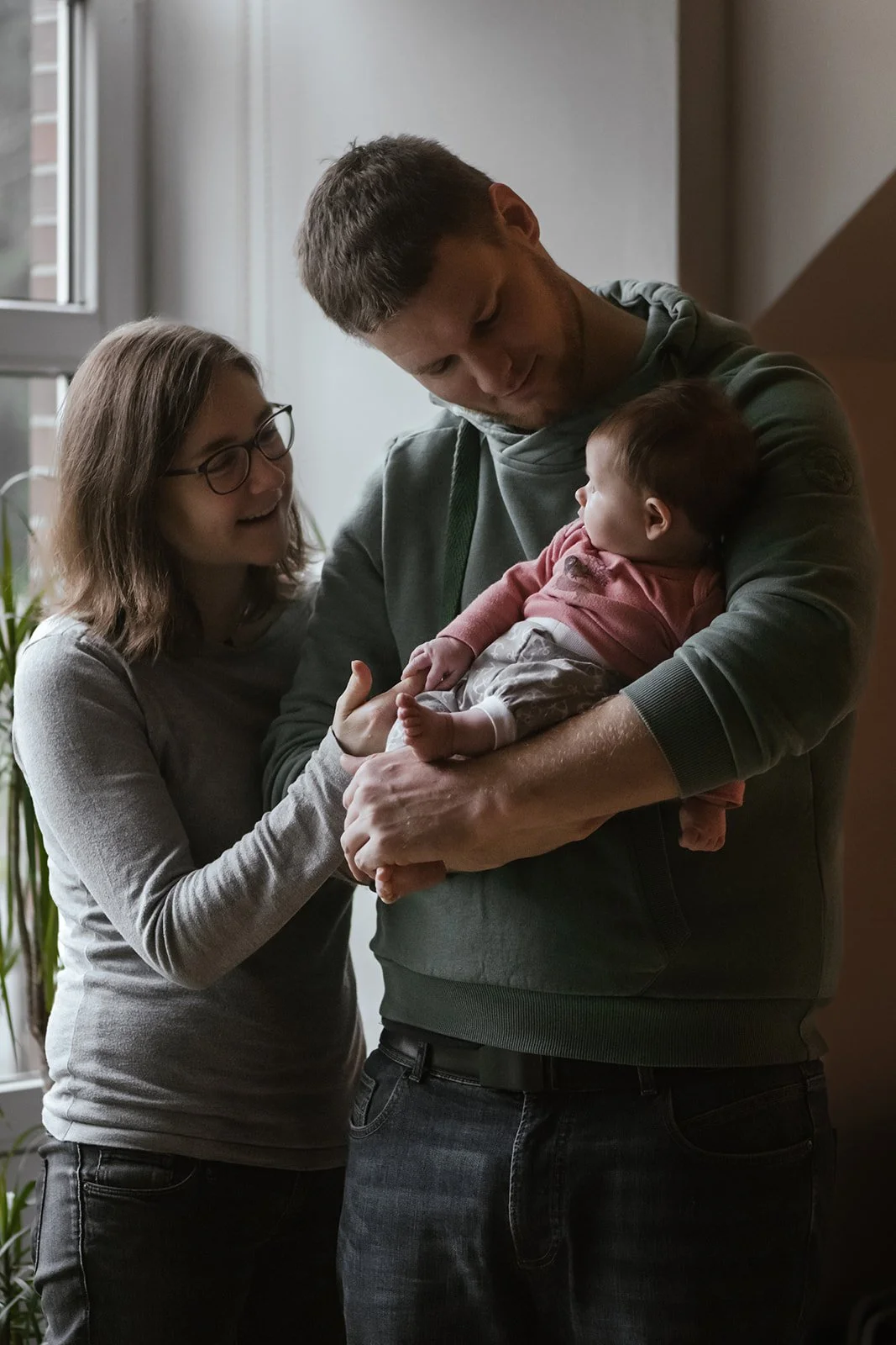 Ein junges Paar hält eine schlafende Baby im Arm, während die Frau lächelnd zuschaut.