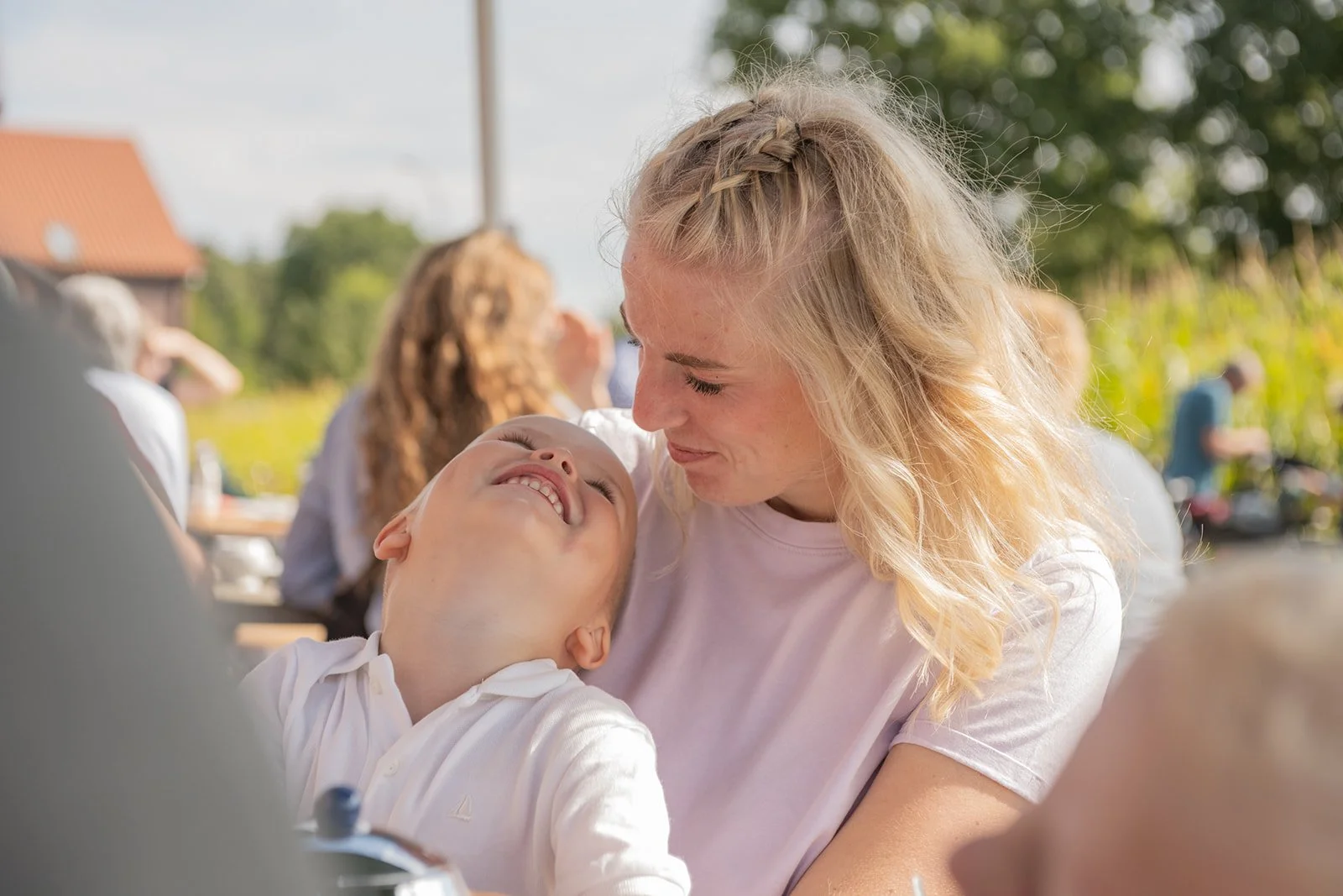 Eine lachende Frau mit blonden Haaren trägt ein Kind, das überglücklich ist, bei einem Outdoor-Essen an einem sonnigen Tag.