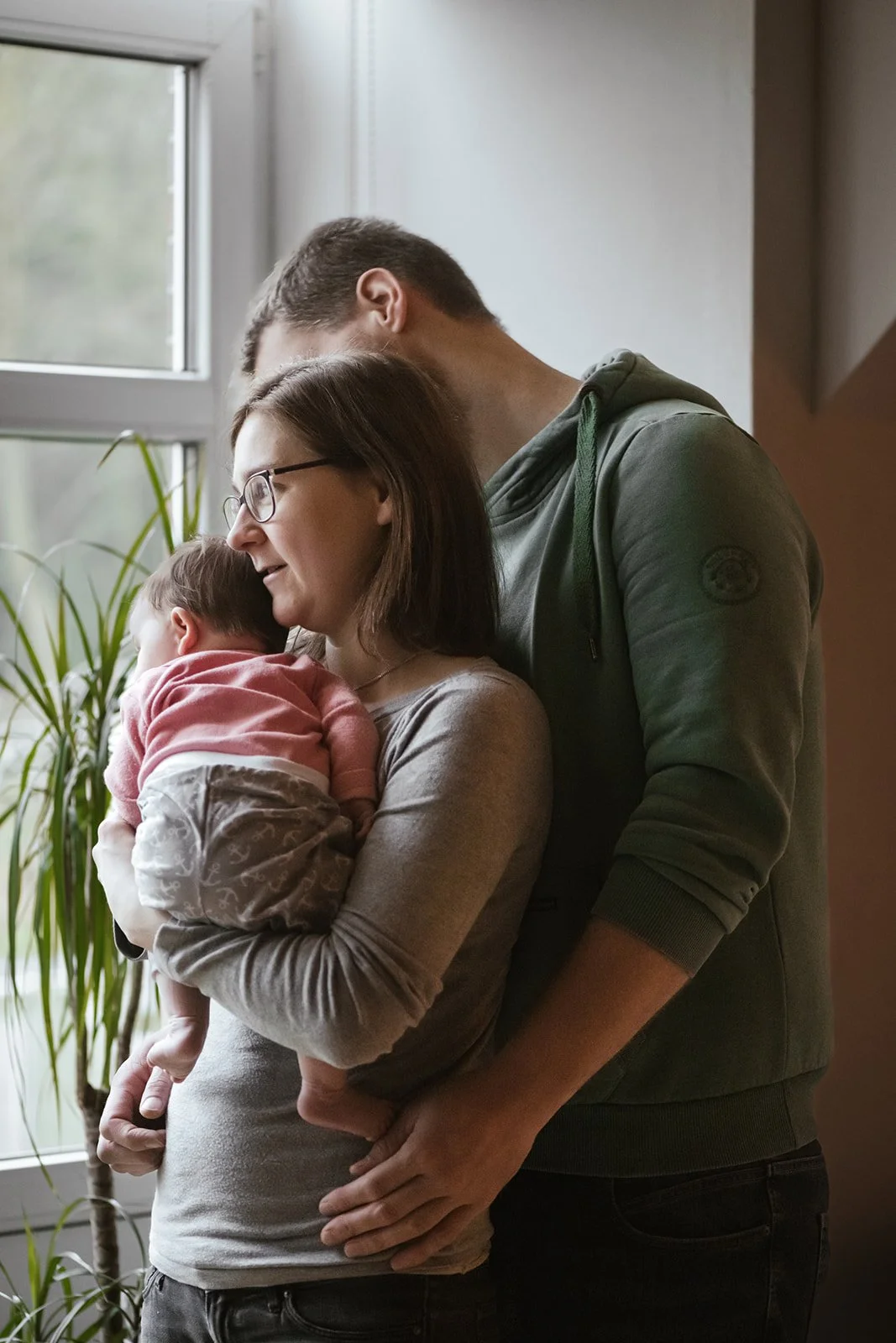 Ein Elternpaar hält ein schlafendes Baby in ihren Armen, vor einem Fenster mit grünem Zimmerpflanze im Hintergrund. Homestory in Ostfriesland