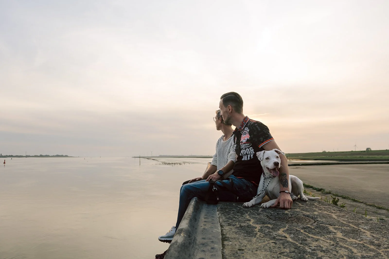 Zwei Menschen und ein Hund sitzen am Ufer eines Flusses bei Sonnenuntergang, die Landschaft ist flach und weit, mit einigen Windkraftanlagen im Hintergrund.