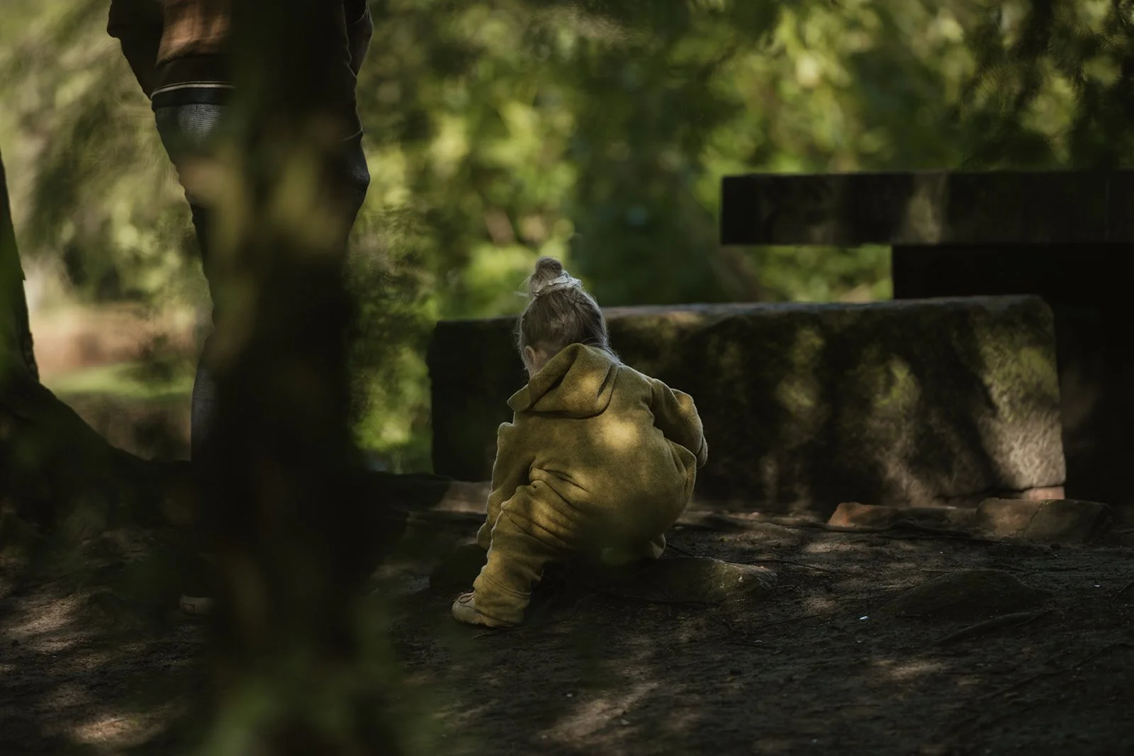 Ein junges Mädchen in einem braunen Kapuzenpullover sitzt auf dem Boden, während jemand an ihrer Seite im Wald steht, umgeben von grünen Bäumen und Schatten.