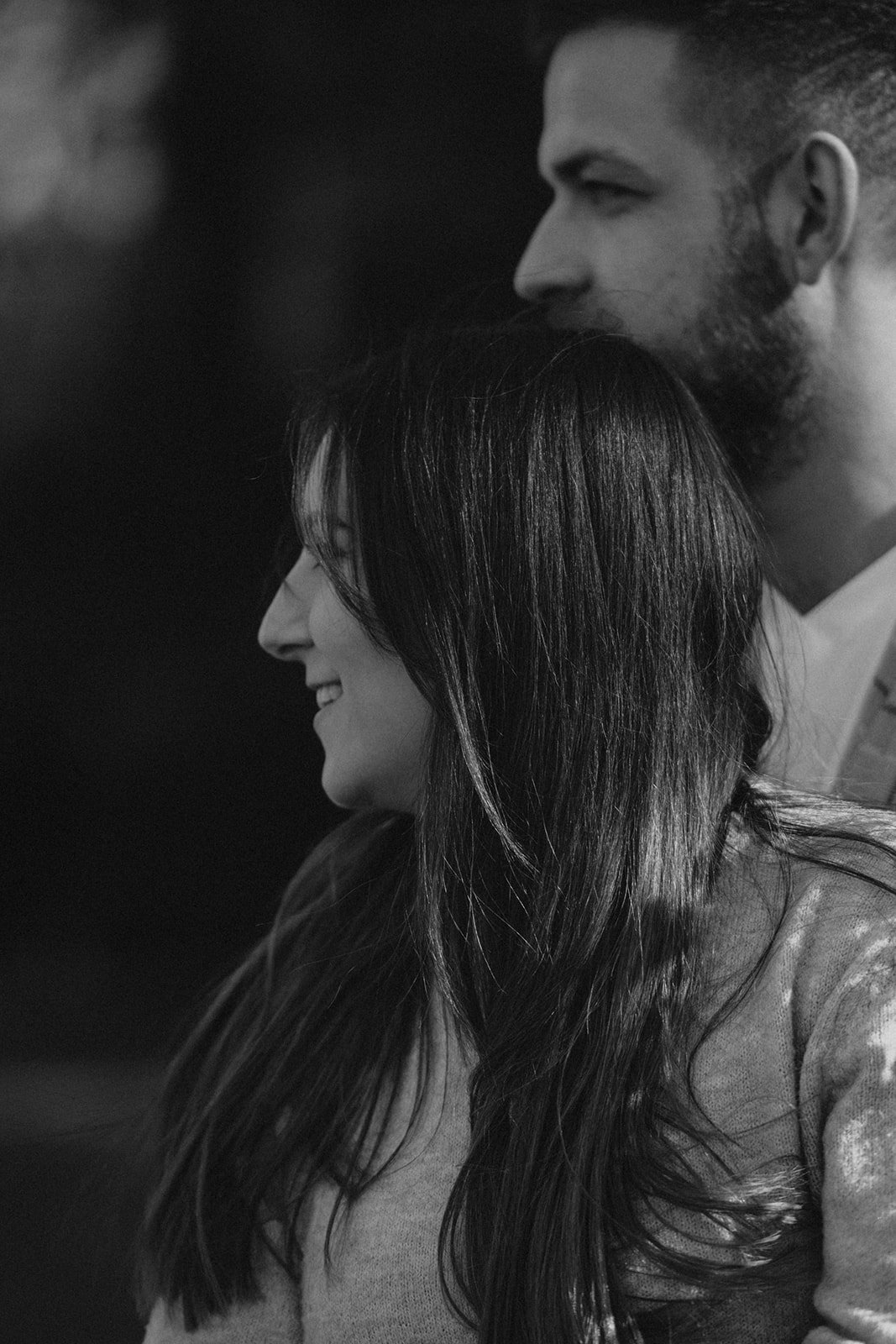 Ein schwarz-weiß Foto eines lachenden Paares, im Profil sichtbar, vor einem dunklen Hintergrund. Verlobung.