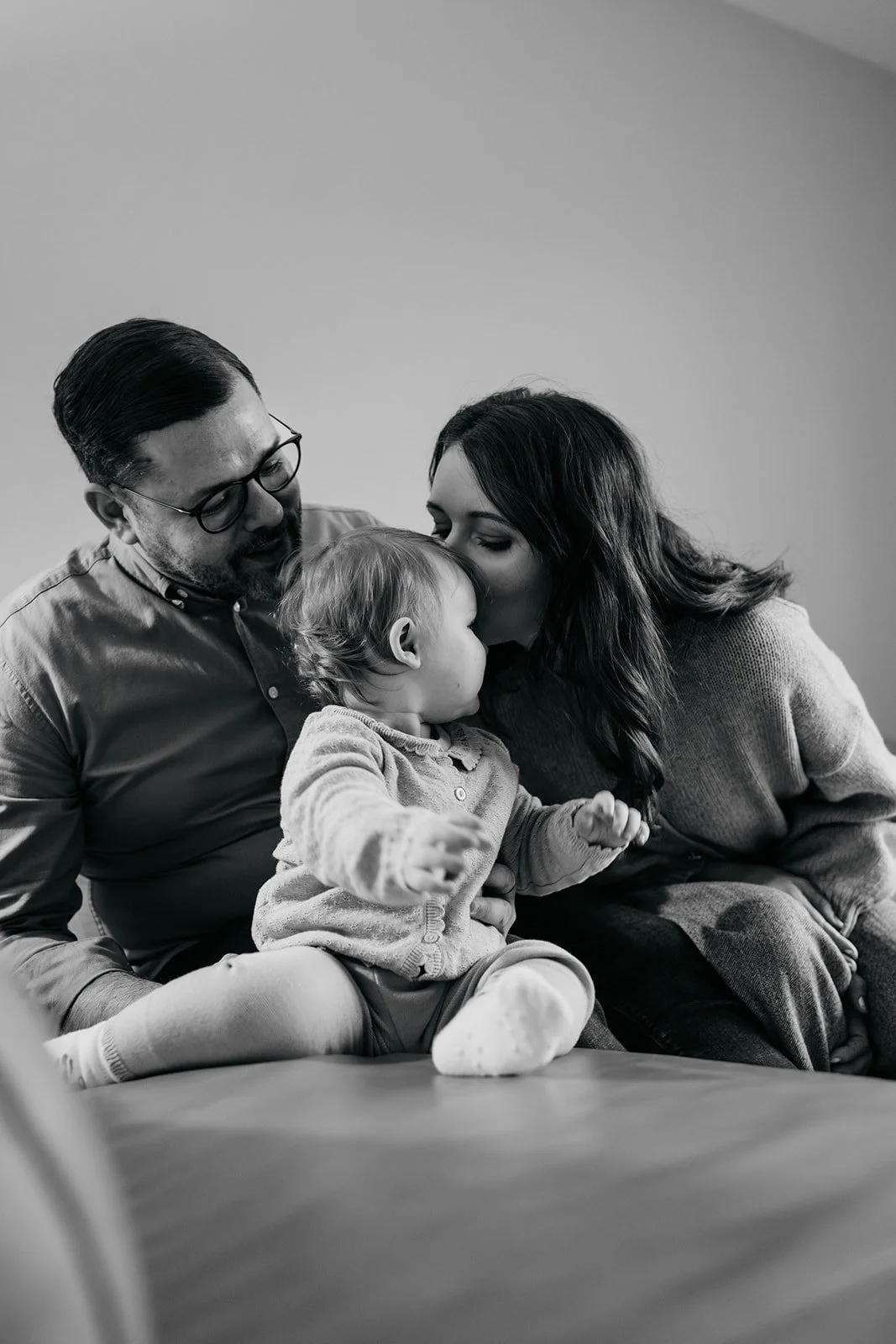 Eine Familie bestehend aus einem Mann, einer Frau und einem kleinen Kind, die eng zusammen sitzen. Der Mann trägt Brille und hat dunkles Haar, die Frau hat lange dunkle Haare. Das Kind sitzt auf einem Tisch und trägt einen Pullover. Die Familie teilt